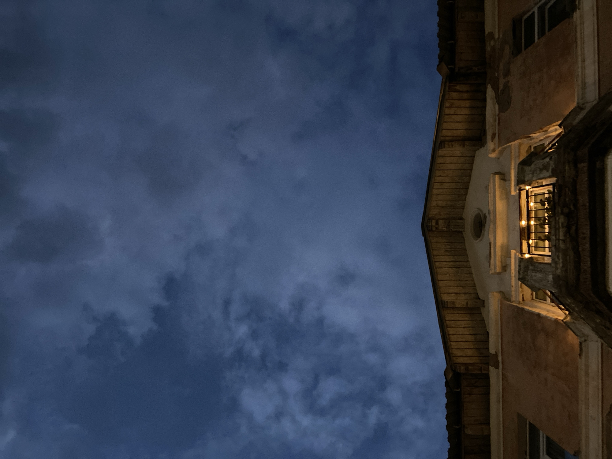 The image captures a low-angle view of the upper facade of a building against a dark, cloudy sky. The building appears to be old, with weathered peach-colored walls and wooden elements under the eaves. A small balcony with a black metal railing is visible, illuminated by warm, yellow lights, possibly from candles or string lights, suggesting it's evening or night. The sky is filled with dark, textured clouds, indicating an overcast or potentially stormy condition. There are no people or animals visible in the frame. The architecture suggests a residential or older commercial building. No specific details are provided to place this image in Votkinsk, Russia; the description is based solely on the visual content of the photograph.