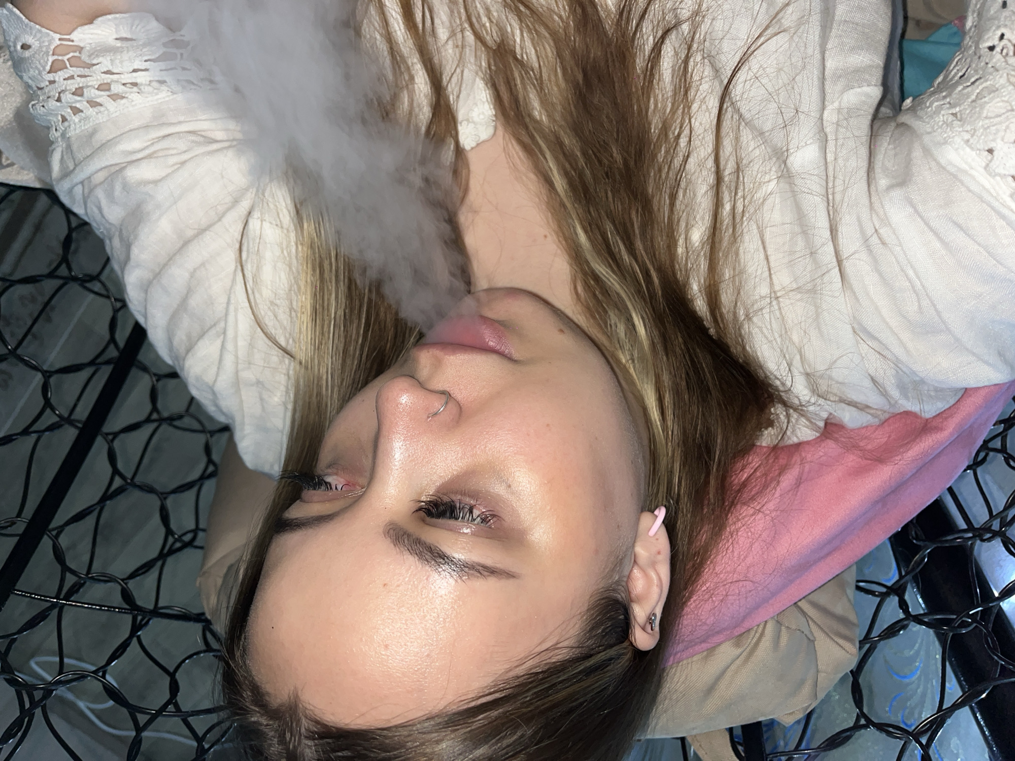 A young woman with light skin, light brown hair, and a nose ring is lying on what appears to be a bed or surface with a black wire frame. She is wearing a white, lacy-trimmed shirt. There is a cloud of vapor or smoke emanating from her mouth. A pink pillow is situated near her head. The location is not discernible.