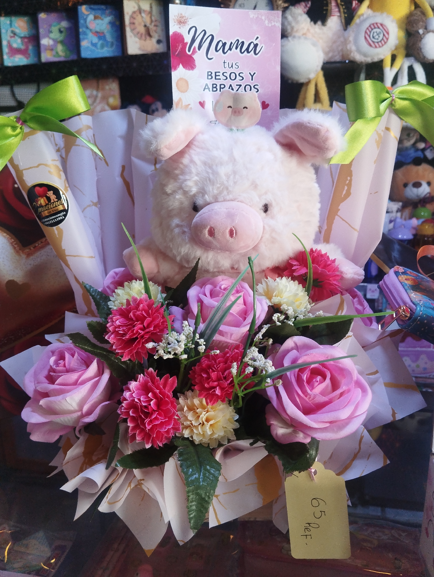 In Pampatar, Venezuela, a floral arrangement featuring a light pink stuffed pig is displayed. The pig sits atop a bouquet of pink roses, small red carnations, and white baby's breath. A card with the Spanish phrase "Mamá tus besos y abrazos" (Mom, your kisses and hugs) is positioned behind the pig. The arrangement is wrapped in patterned white and gold paper tied with green ribbon. A yellow tag with "65 Ref." written on it is attached. The arrangement is in a shop setting, with shelves of various gifts and toys visible in the background.