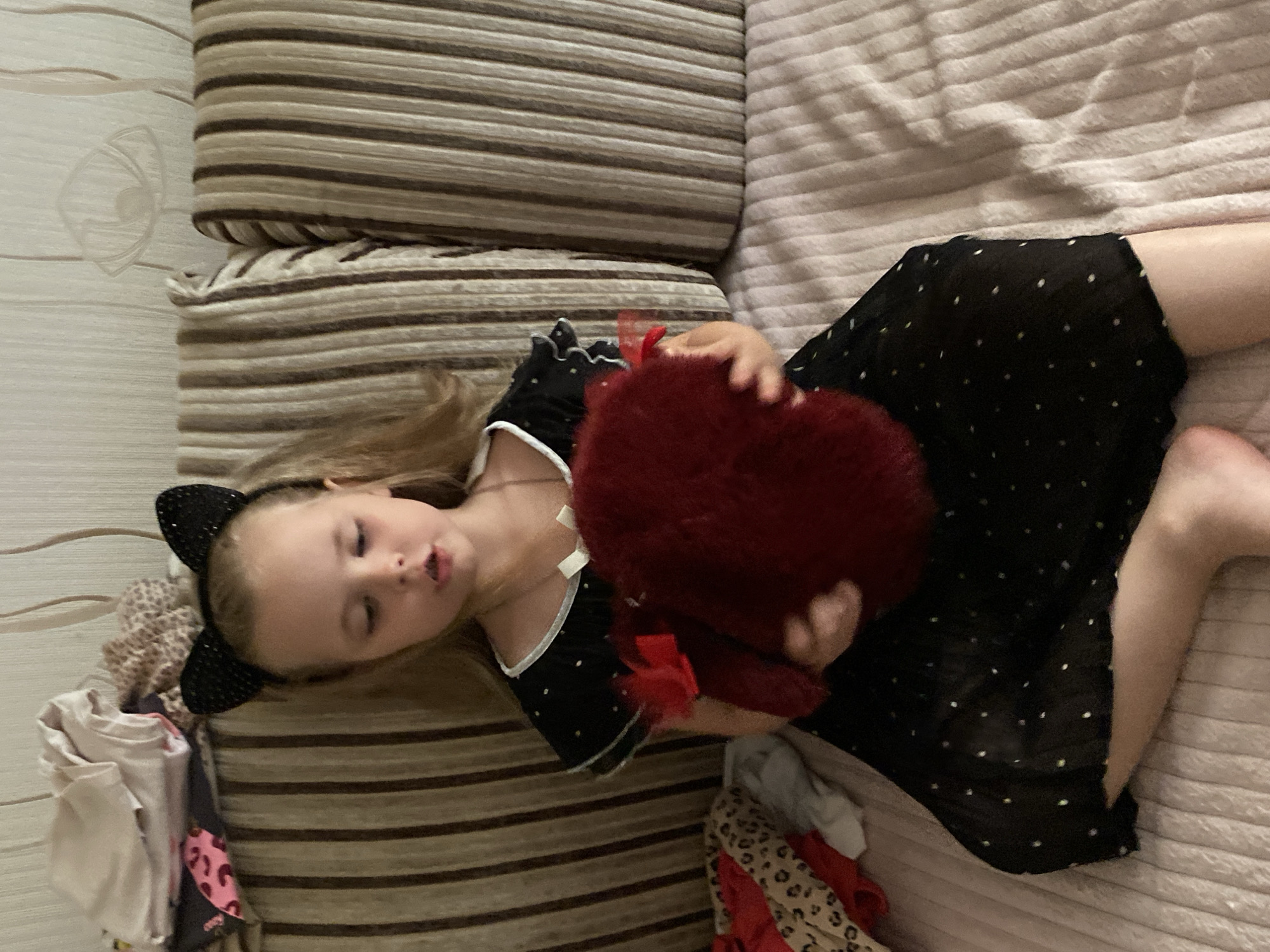 A young girl with blonde hair and black cat ears is lying on a striped couch. She is wearing a black dress with white polka dots and has a red, heart-shaped fuzzy object in her hands. Her eyes are closed, and her mouth is slightly open, suggesting she is sleeping or resting. Several pieces of clothing are scattered around her on the couch and on the floor next to it, including a pink and black leopard print garment. The setting appears to be indoors, likely a living room, with a textured, pale pink blanket covering part of the couch. The lighting suggests it could be daytime, possibly late afternoon or evening, as there are shadows present. The overall mood is relaxed and intimate.