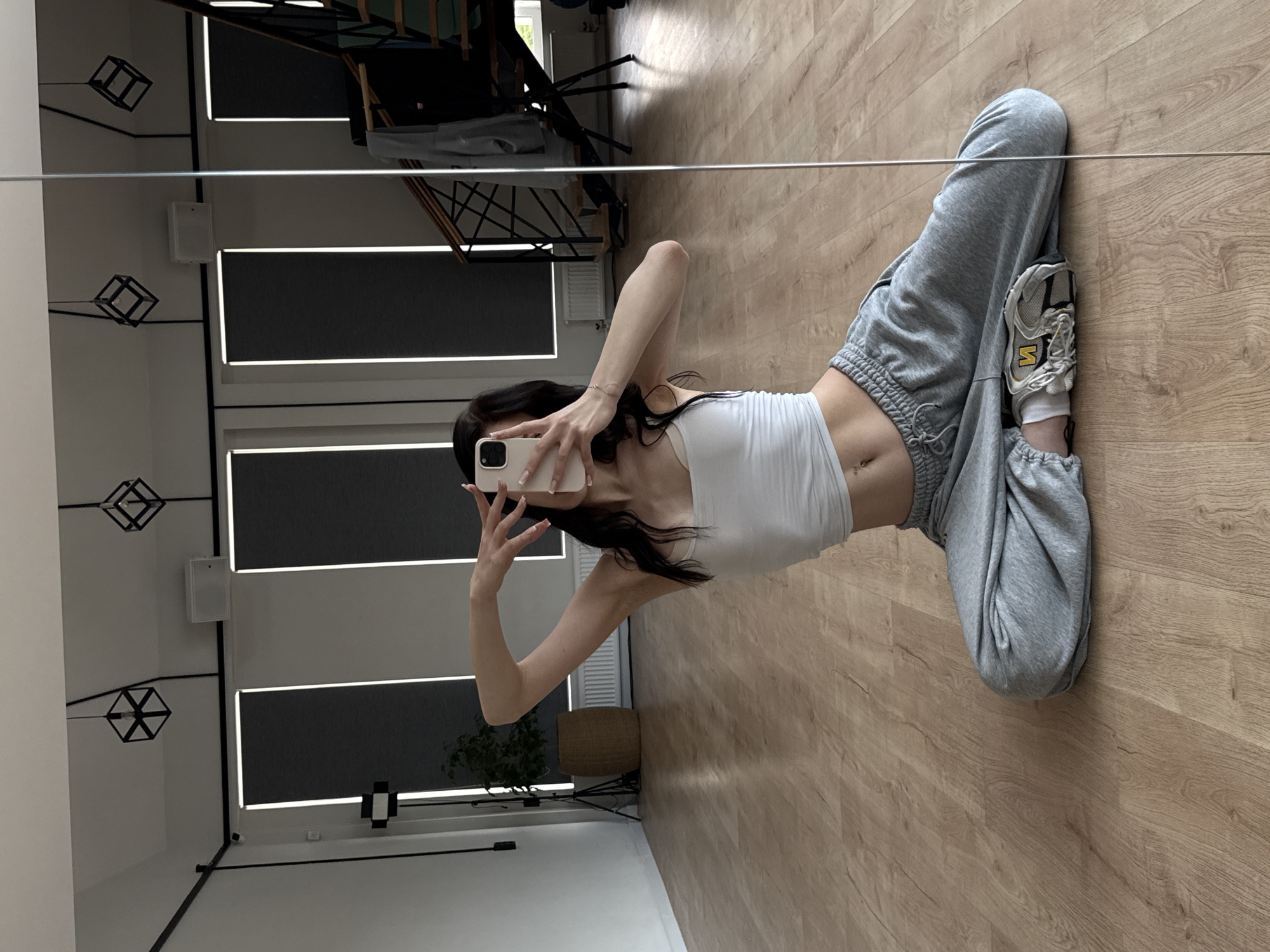 A person is sitting on a wooden floor, facing a mirror. The person is wearing a white crop top and gray sweatpants. Their right arm is raised, and their hand is near their face, holding a mobile phone. The phone is being used to take a selfie. The background shows a room with large windows covered by dark blinds, a plant in a woven basket, and industrial-style hanging lights. A staircase is visible in the reflection of the mirror, along with some clothing items. The location is identified as Agalarov Estate KP, Russia, suggesting this might be a residential or studio space within that area. The lighting appears to be natural, coming from the windows.