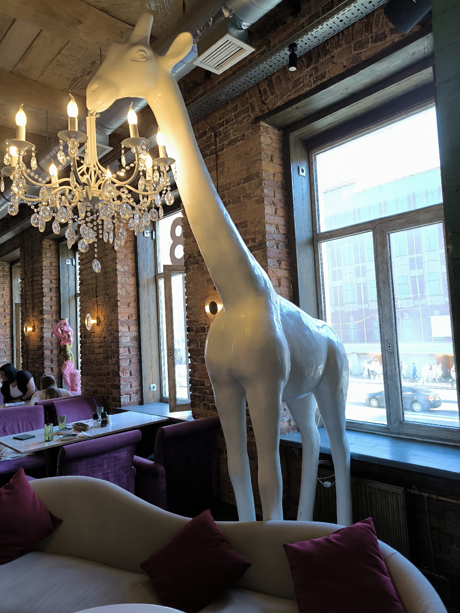 This image presents a detailed indoor scene of a commercial establishment, identified as a cafe or restaurant in Saint Petersburg, Russia.
The primary subject is a large, smooth, white full-body statue of a giraffe, positioned on the right side of the frame, extending from the floor towards the ceiling. Its head is angled slightly to the left, towards a prominently featured white chandelier in the upper left quadrant. This chandelier is ornate, with multiple arms, several lit candle-shaped bulbs, and numerous clear crystal pendants.
The interior architecture features exposed red brick walls and pillars throughout the space. The ceiling consists of wooden planks, with visible metallic ductwork and perforated cable trays running across it, equipped with small black track lights. Warm-toned circular wall sconces are mounted on the brick pillars.
In the mid-left, two women are seated at tables featuring white tabletops and purple velvet upholstered chairs or booth seating. One woman, with dark hair, is seen in profile, looking down. Green glasses and small bottles are visible on the tables. Further back on the left, against a brick pillar, a pink, draped female figure, resembling a mannequin, stands.
Large, multi-pane windows with dark wooden frames form the right side of the room, offering an exterior view of a city street during daylight hours. Through these windows, pedestrians are visible walking on a sidewalk, and a dark sedan is parked on the street. Buildings across the street feature large glass facades, reflecting ambient light. A black horizontal radiator is situated below the window. An open glass door, integrated into the window structure, is also visible further back on the left, leading to the street.
In the immediate foreground, a cream-colored, curved sofa is present, furnished with two dark red cushions.