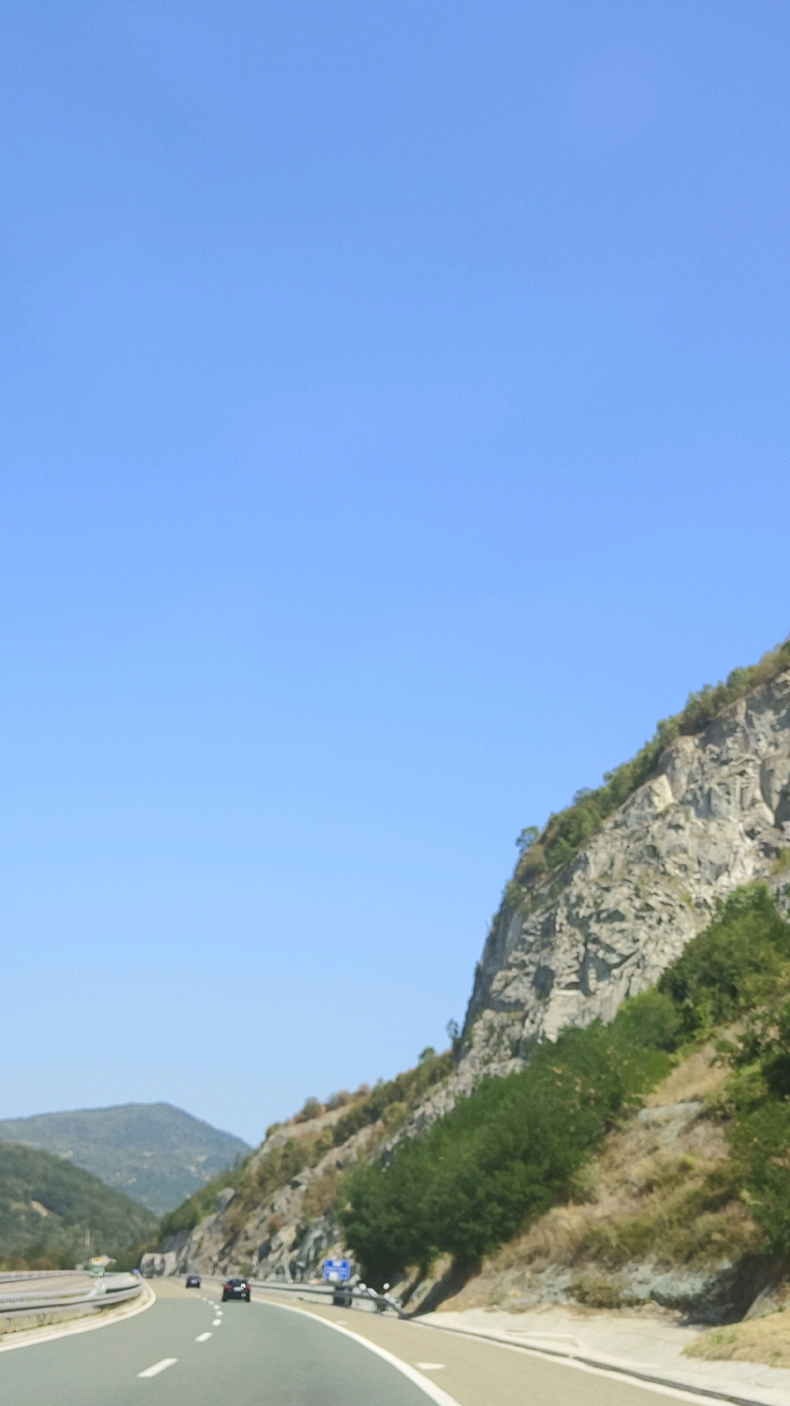 The image captures a view from a vehicle traveling on a highway in the Vladicin Han Municipality, Serbia. The highway curves to the right, with two lanes visible in the foreground. The road is paved and marked with white lines. On the right side of the highway, a steep, rocky mountainside rises sharply, covered in sparse vegetation and trees. The mountain is gray and textured, indicating exposed rock faces. On the left side of the highway, rolling green hills extend into the distance. Several cars are visible on the road, including one dark-colored car in the right lane and another dark car further ahead in the left lane. Blue road signs are present on the right side of the highway. The sky is clear and bright blue, suggesting a sunny day. The overall scene depicts a mountainous highway landscape.