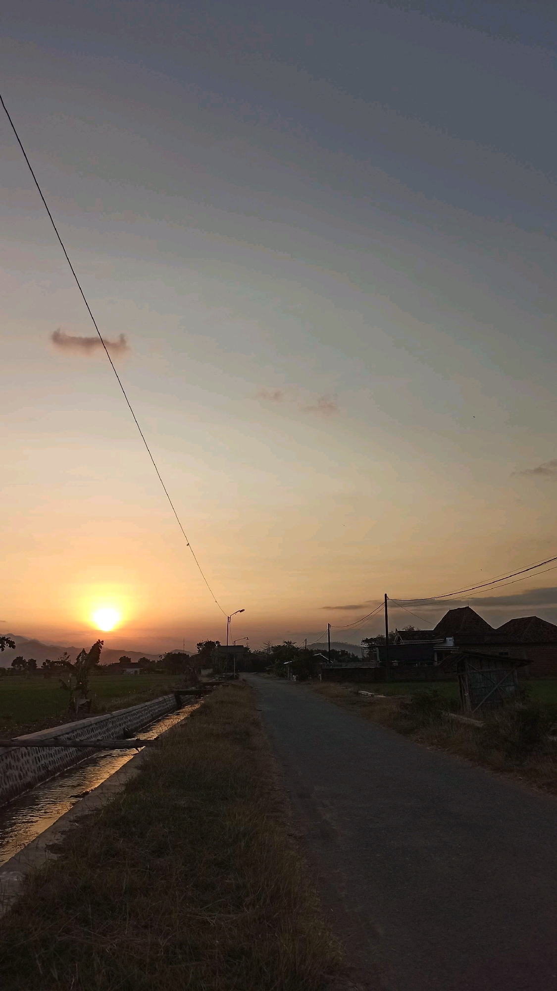 An outdoor, vertical, medium-shot photograph captures a serene rural landscape at dusk in Josari, Indonesia. The sky transitions from a soft, warm orange near the horizon to a cool, pale blue at the top, with a few wispy clouds scattered across. The sun, a bright orange orb, is setting behind distant mountains, casting a warm glow over the scene. A prominent power line stretches diagonally across the sky, anchored by a pole on the right side of the frame.
In the foreground, a paved road runs from the bottom center of the image towards the horizon, flanked by dry, tall grass on the right. To the left of the road, a concrete irrigation channel filled with reflecting water runs parallel to the road. Beyond the channel, a green rice paddy is visible, dotted with trees and some small buildings. In the distance, several houses with reddish-brown tiled roofs are clustered, along with more vegetation and rolling hills. The overall atmosphere is peaceful and tranquil, typical of a rural sunset. There are no people or animals visible in the frame.