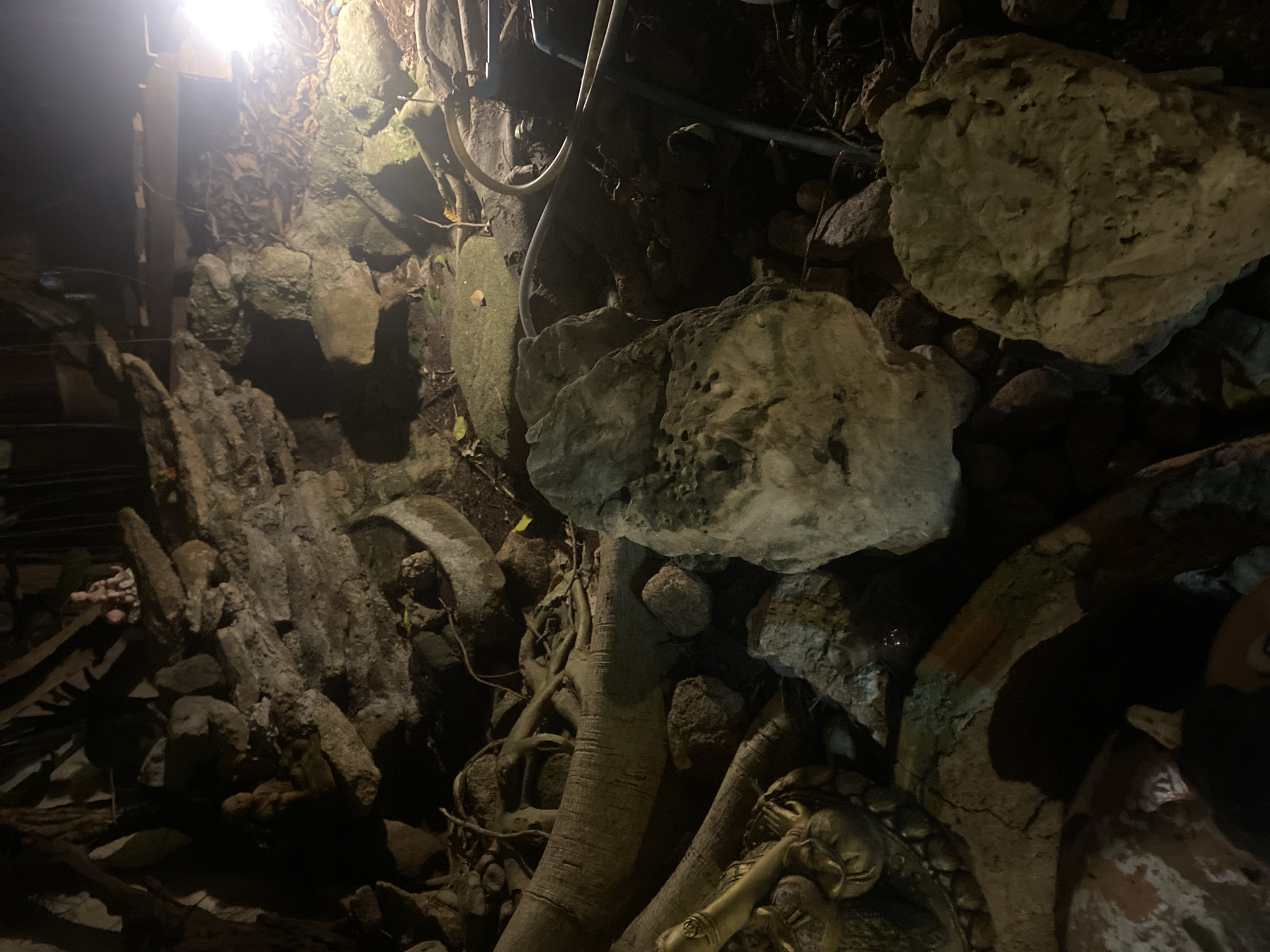 This image shows a scene with rocks, tree roots, and what appears to be part of a statue, likely located in Baan Don Sai, Thailand. A light source illuminates the upper left area, casting shadows throughout. The environment consists of stacked rocks and intertwined tree roots, with a piece of a golden statue visible in the lower right. There are no people visible in the frame.