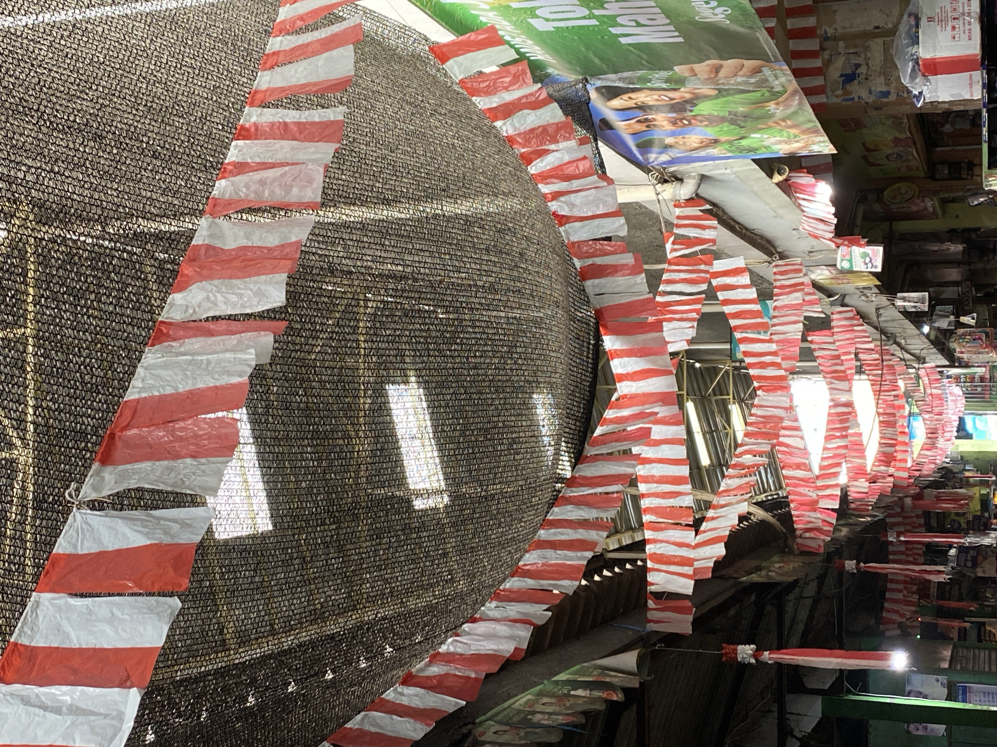 This is a photograph taken from a low angle looking upwards into a covered market in Ambarketawang, Indonesia. Red and white striped flags are strung across the market, creating a festive or patriotic atmosphere. The flags are made of a thin material and appear to be attached to strings or wires that are hung from the dark, net-like covering that forms the ceiling of the market. This covering is supported by structures that are partially visible, including wooden beams and possibly metal framing.
The market stalls below are not clearly visible in detail, but there are hints of merchandise and signage. To the right of the frame, a large green banner with text and an image of people holding Sprite bottles is prominently displayed. Further back, various colorful signs and products can be glimpsed. Hanging decorations, also featuring red and white elements, are visible in the foreground, suggesting a celebratory occasion, possibly related to Indonesian Independence Day given the colors.
The lighting suggests it is daytime, with natural light filtering through gaps in the market's roof and illuminated by artificial lights within the market. The overall impression is of a bustling, covered marketplace decorated for an event.