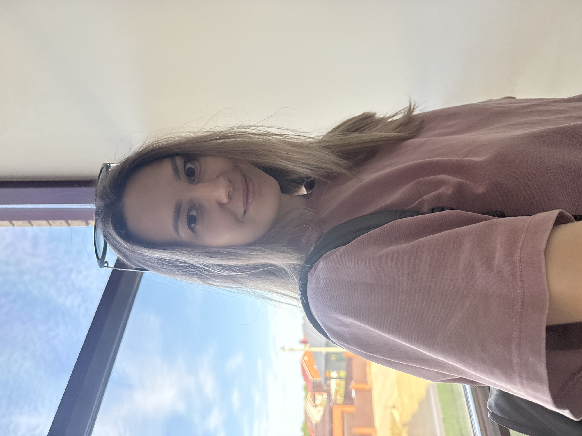 A young woman with shoulder-length, blonde hair is posing for a selfie near a window. She wears a rose-colored shirt, and sunglasses are perched atop her head. Outside the window, a partial view shows construction equipment and buildings, consistent with an urban environment, possibly in Noviy, Russia. Blue skies are visible through the window. The subject's expression is cheerful, and she is looking directly at the camera. A dark backpack strap is also visible.
