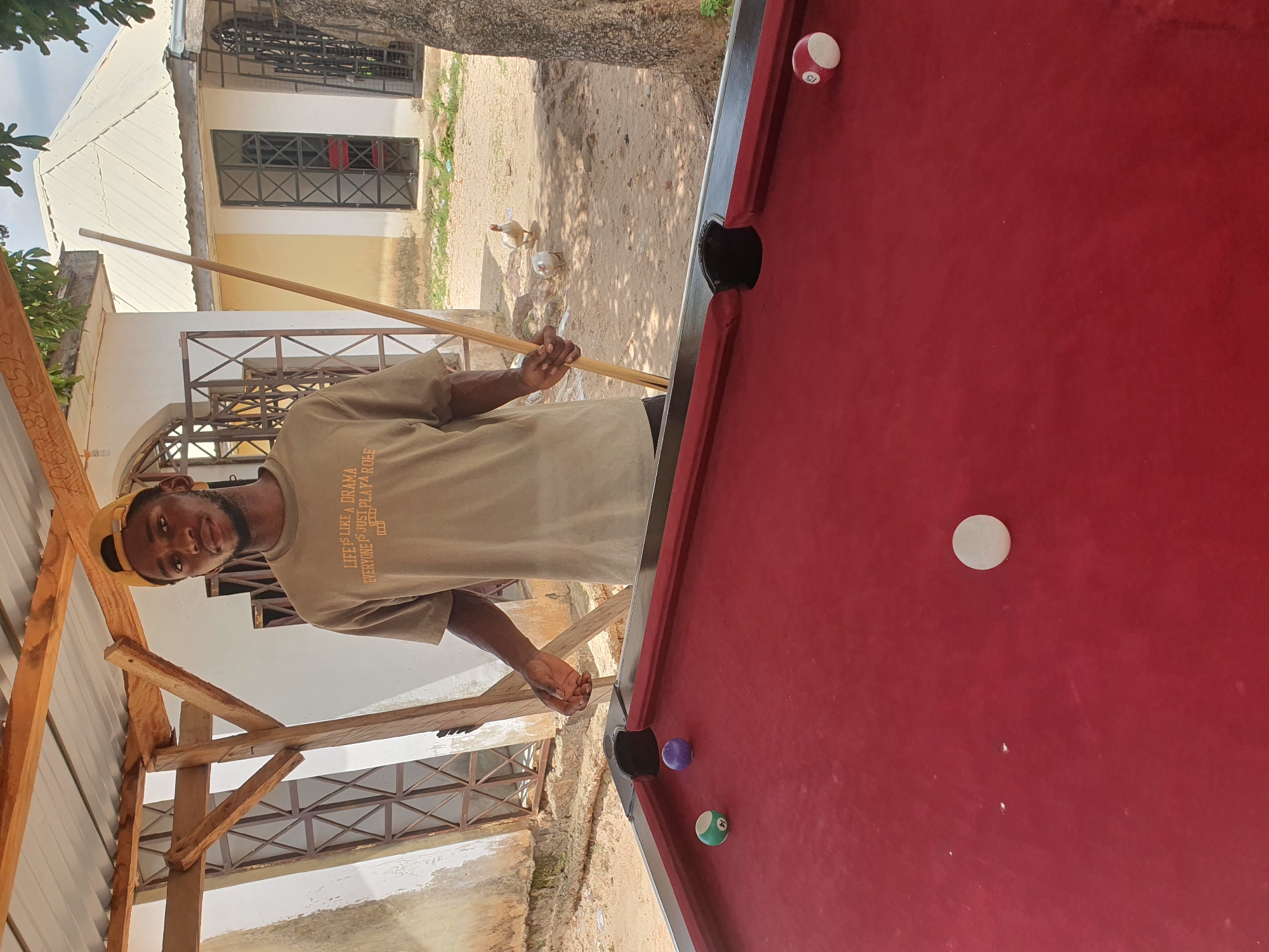 A young Black man wearing a beige t-shirt and a yellow baseball cap is standing next to a red billiard table. He is holding a pool cue, appearing to be in the process of playing. The t-shirt has text on it that reads, "LIFE IS LIKE A DRAMA EVERYONE IS JUST PLAYING THEIR ROLE." He is looking towards the camera with a slight smile.
The scene appears to be outdoors, with a building and some trees in the background. The ground is sandy or dirt. There are a couple of ducks or chickens walking in the background. The billiard table is red with black pockets and has several balls on it. One ball, with the number 15, is red and white. Another ball is green with the number 11. A purple ball is also visible. A white cue ball is in the center of the table. The lighting suggests it is daytime, likely a sunny day given the shadows. The setting seems to be a casual, perhaps residential, area in Lokoja, Nigeria. There are wooden structures and a corrugated metal roof visible to the left of the man.