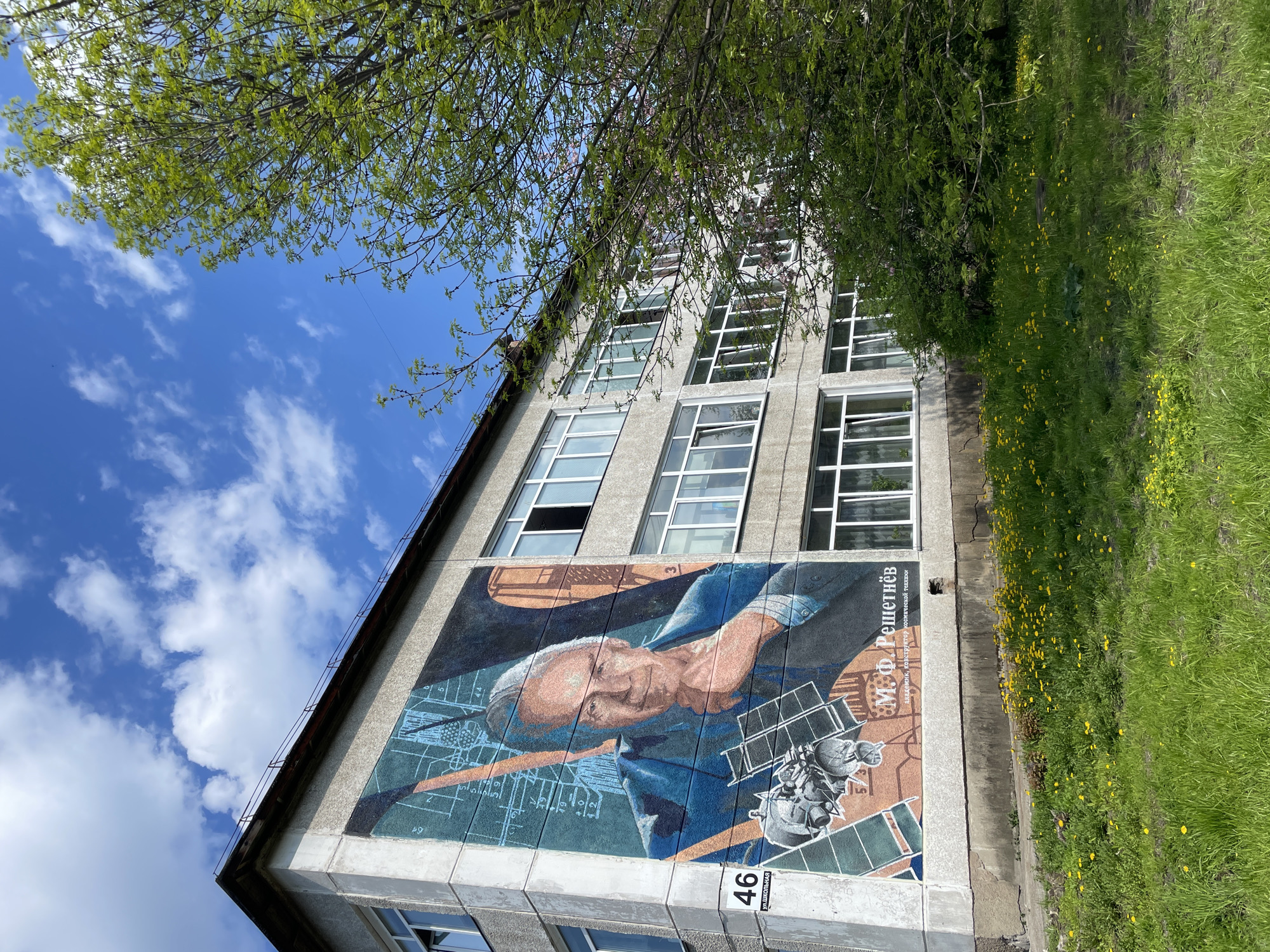The image shows a building with a large mural of M.F. Reshetnev, a Soviet spacecraft engineer and designer. The mural also features drawings and schematics related to space technology, as well as a satellite design. The building is located at 46 Ulitsa Shkolnaya in Zheleznogorsk, Russia. The building has multiple windows and is surrounded by grass and some greenery. The sky above is blue with white clouds, and there is a tree branch partially obscuring the view of the building.