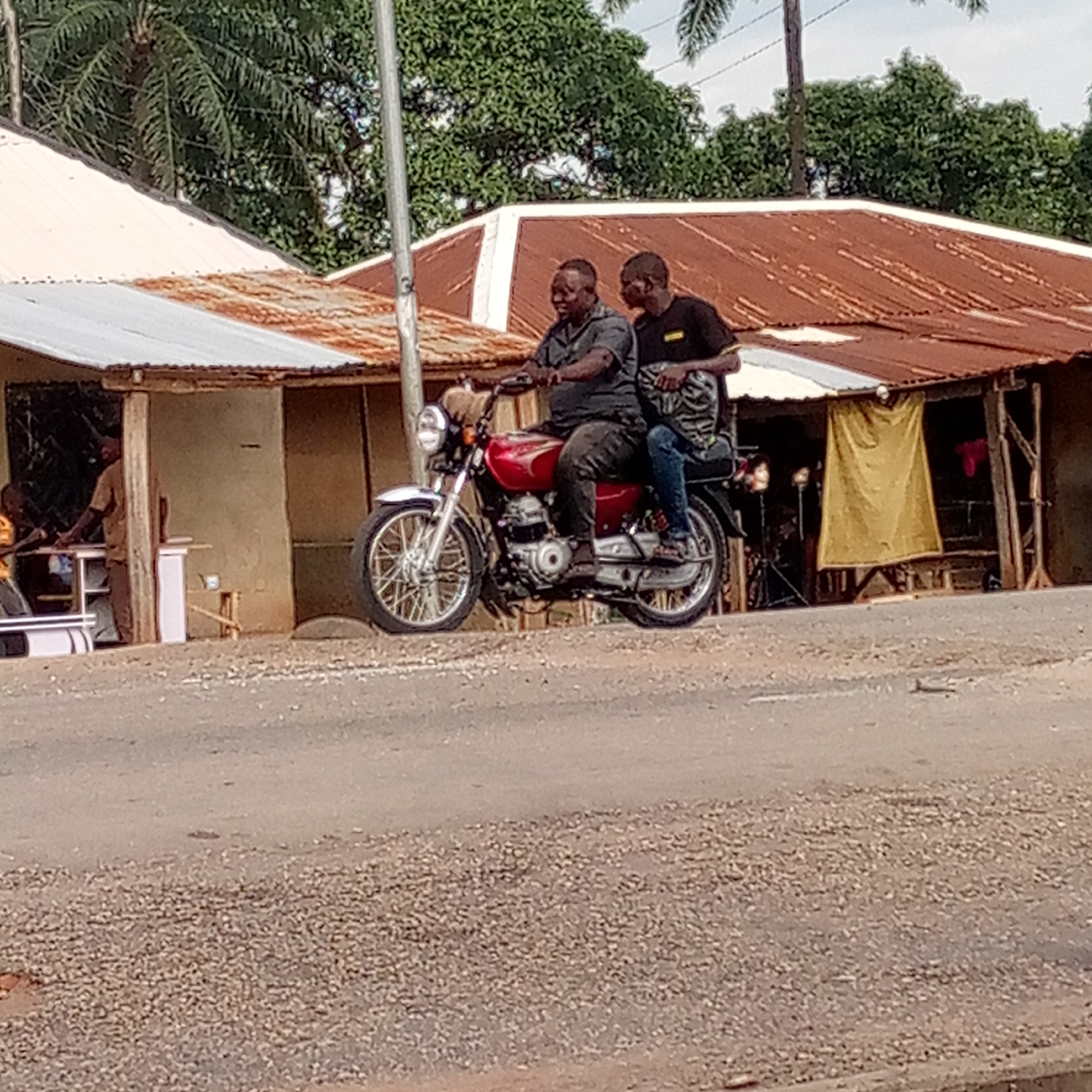 Two men ride a red motorcycle on a paved road in Mararaba, Nigeria. The rider, in a dark grey shirt and pants, sits upright. The passenger, in a black shirt and blue jeans, sits behind the driver with his right hand on the driver's shoulder. The motorcycle's front headlight is on. Two shacks with corrugated metal roofs are in the background; one with a white roof to the left and another with a rusted roof to the right. A few people stand in the shack on the left. A yellow curtain hangs in the shack on the right. Palm trees are visible in the background. The road appears to be paved but in rough condition.
