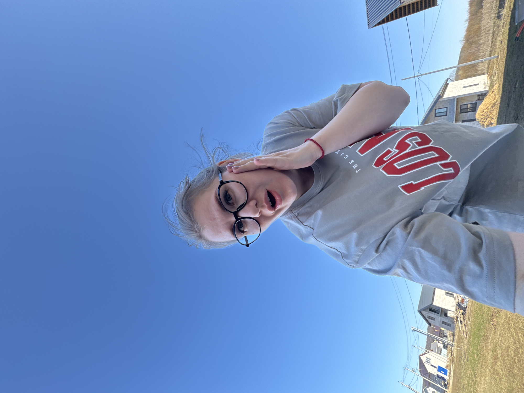 A young woman wearing glasses and a gray t-shirt is taking a photo from a low angle, looking up at her. She is positioned against a bright blue sky. Her hand is raised near her face. The background shows houses and power lines, suggesting a residential area. The location appears to be Gornyi (Dvurechenskoe S/P), Russia, based on visual cues in the image.