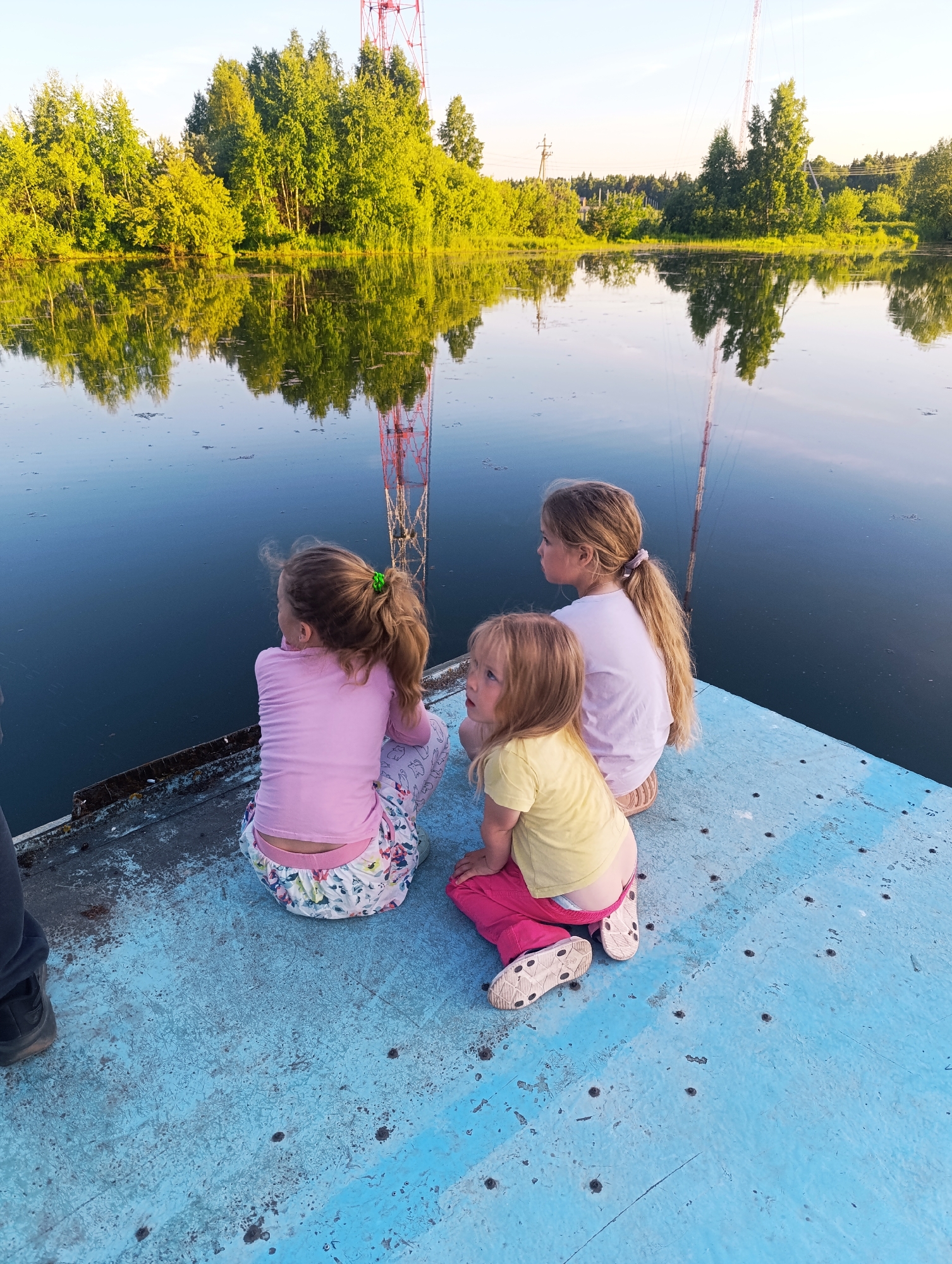 Three young females are seated on a weathered, light blue surface, consistent with a dock or pier, facing a calm body of water. An adult's left foot and lower leg, dressed in dark clothing and a dark shoe, are visible in the extreme lower-left corner. The leftmost girl, with long blonde hair secured by a green tie, wears a pink long-sleeved shirt and white patterned pants, partially revealing a light-colored undergarment above the waistband. The middle girl, possessing long blonde hair in a light-colored ponytail, is clad in a light pink short-sleeved top and light-colored pants. The youngest girl, on the right, has short blonde hair and wears a yellow short-sleeved top with pink pants, also exposing a light-colored undergarment. All three girls are positioned in a crouch, their backs mostly towards the viewer, looking intently towards the water.
The water, appearing dark blue, reflects a dense line of vibrant green trees on the opposite bank under a bright, clear sky. A tall, red and white lattice communication tower is visible on the right side of the background, its reflection clearly depicted in the water. Scattered dark particulate matter floats on the water's surface. The light blue surface on which the individuals are seated exhibits significant wear, with chipped paint, dirt, and numerous small, dark, circular indentations or marks. The scene is illuminated by natural sunlight, suggesting daylight hours. The specific location is not identifiable within the image, occurring in Unknown, Unknown.