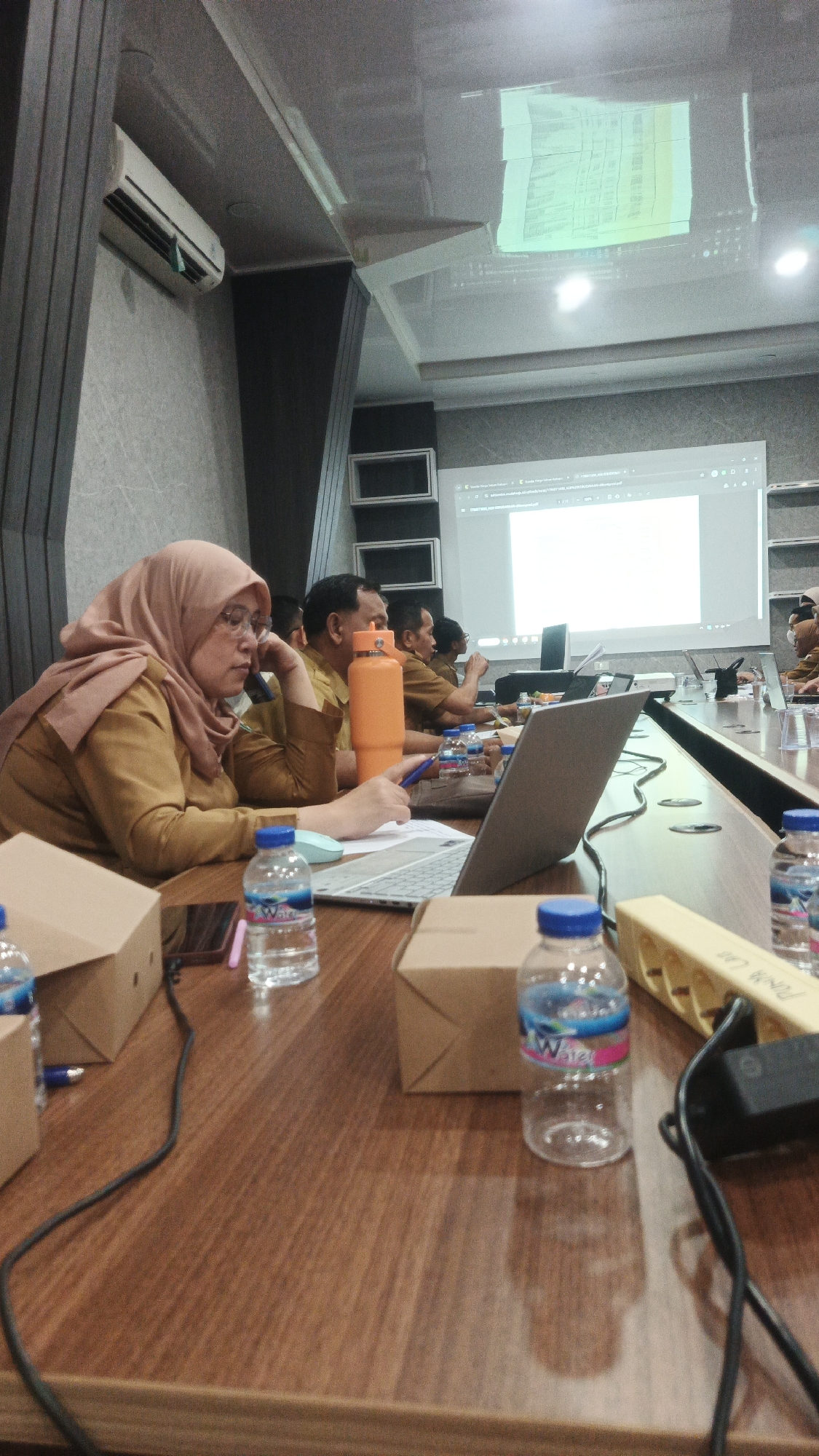 The image depicts an indoor meeting or presentation scenario, likely within an office or conference room. In the foreground, a wooden conference table is laden with various objects. A woman wearing a light brown hijab and a mustard-colored outfit is seated at the table, appearing to be engaged with a laptop and holding a pen. In front of her is a laptop, a blue pen, and a bottle of water. To her right, another bottle of water is visible.
Further down the table, several other individuals are seated, also appearing to be participants in the meeting, though their faces are not clearly visible or are partially obscured. Some are wearing similar mustard-colored attire, suggesting they may be colleagues or part of the same organization. An orange water bottle is prominent among the items on the table.
In the background, a large projection screen displays a presentation, with some text and graphics visible. The room's lighting appears to be a mix of ambient and artificial light, possibly from ceiling fixtures and the projector. An air conditioning unit is mounted on the wall to the left of the frame.
The overall impression is of a professional gathering, possibly a training session, workshop, or regular business meeting. The environment seems functional and equipped for presentations and collaborative work. The location is not specified in the image, but the request mentions "Sukadana, Indonesia" as a potential context for the city. Without further information or visual cues specific to Sukadana, a definitive location cannot be confirmed. However, if this scene is indeed in Sukadana, it would represent a modern meeting space within that Indonesian city.