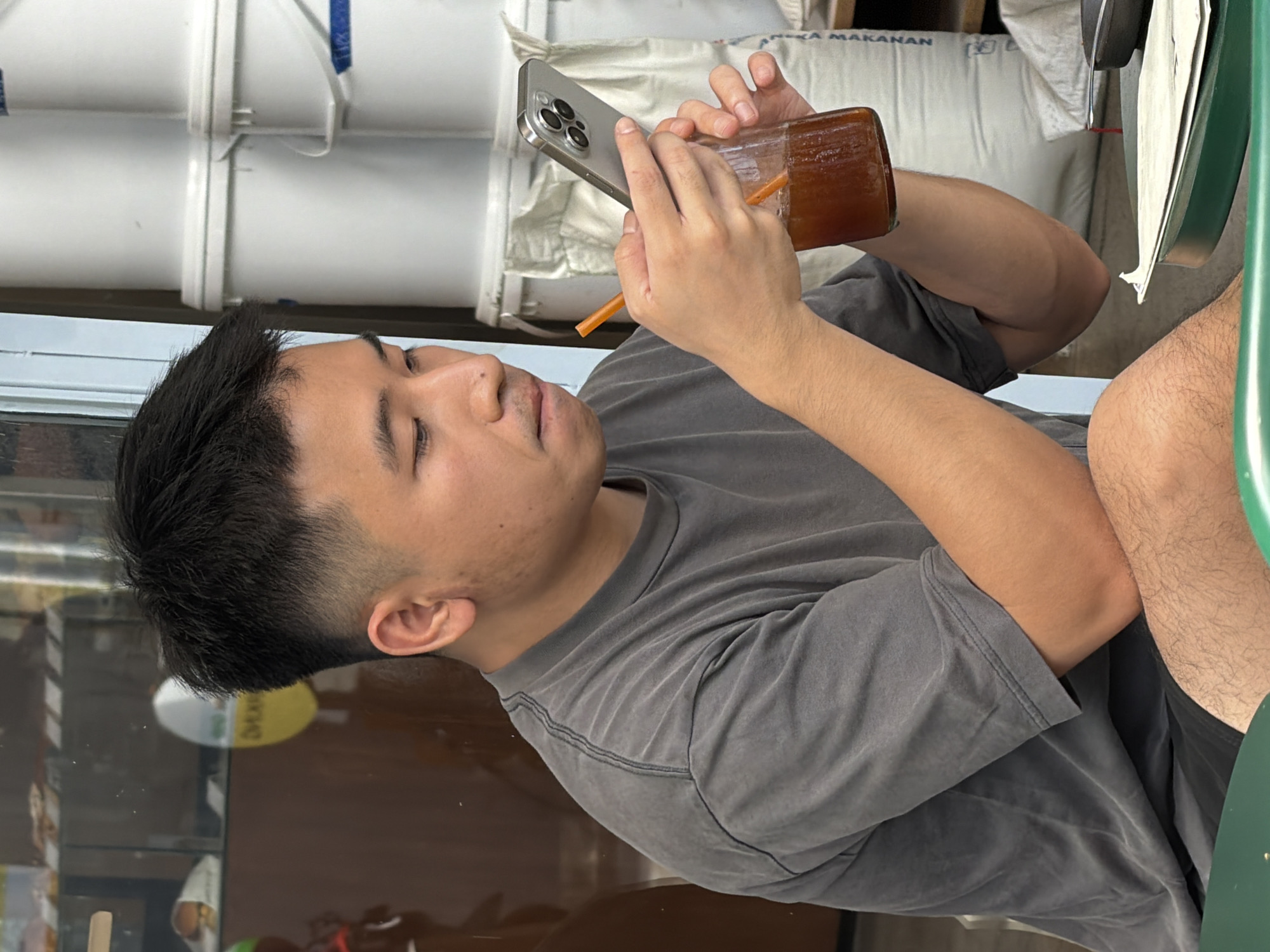 A young East Asian male is seated outdoors, engrossed in his smartphone while holding a glass bottle of iced beverage with a straw. He is wearing a grey t-shirt and dark shorts. The background features stacked white containers, possibly industrial supplies, and a reflective glass facade of a building, suggesting a commercial area. The scene is illuminated by natural light. The location is identified as North Kuta, Indonesia.