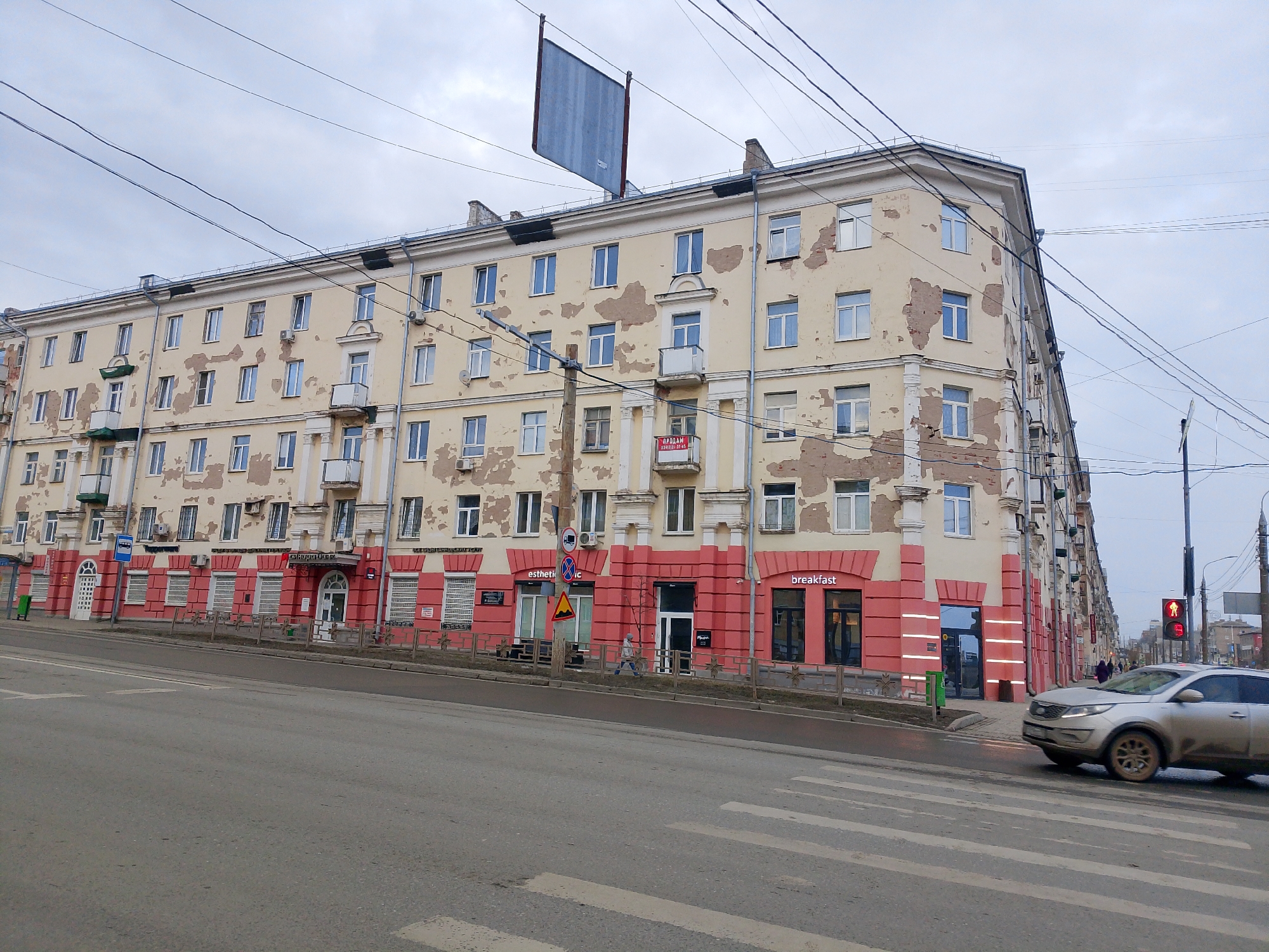 The image shows a five-story building in Izhevsk, Russia, located at the intersection of a wide, gray asphalt street with a marked pedestrian crossing. The building's façade is yellow with peeling paint, and the lower level has a pink facade containing commercial spaces, including one business advertising "breakfast" in English. Power lines crisscross the sky. A large, rectangular sign structure is attached to the building's roof. A silver car is driving through the intersection, and the traffic light is red. A few pedestrians are present in the scene. The sky is overcast, suggesting a cloudy day.