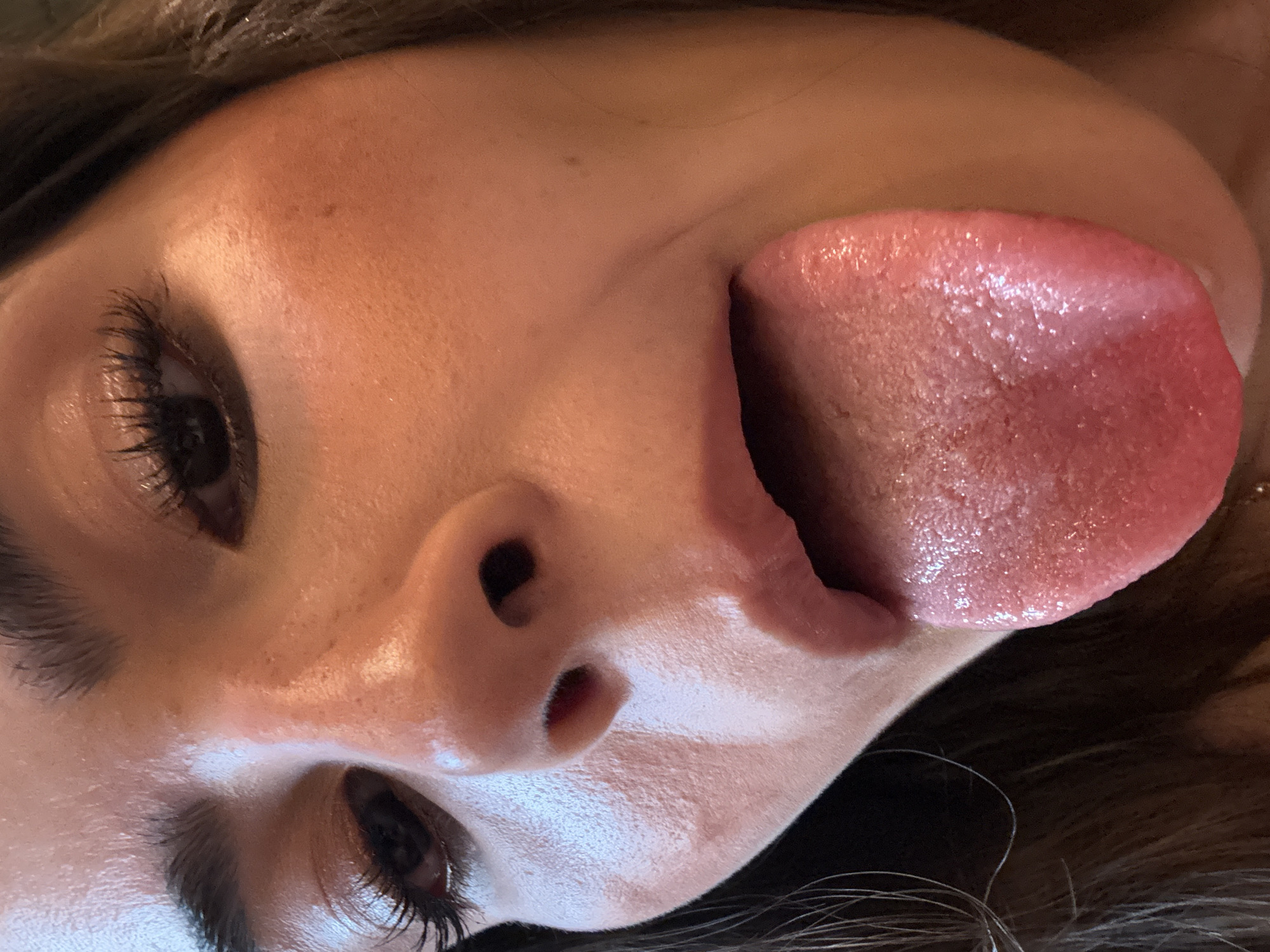 A close-up, low-angle shot features a person's face, with their tongue prominently extended out of their mouth. The person has dark eyes with long eyelashes. The skin appears smooth and possibly slightly damp. The photograph is taken at a close range, capturing intricate details of the face, particularly the texture of the tongue. There is no evidence to indicate this image was taken in San Bernardino Tlaxcalancingo, Mexico.