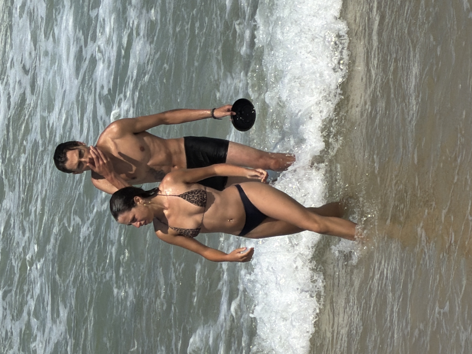 A man and a woman are standing in the water at a beach in Tel Aviv, Israel. The man is wearing black swim trunks and holding a black disc in his right hand. The woman is wearing a leopard print bikini bottom and a black bikini. The water is choppy and the sand is wet.