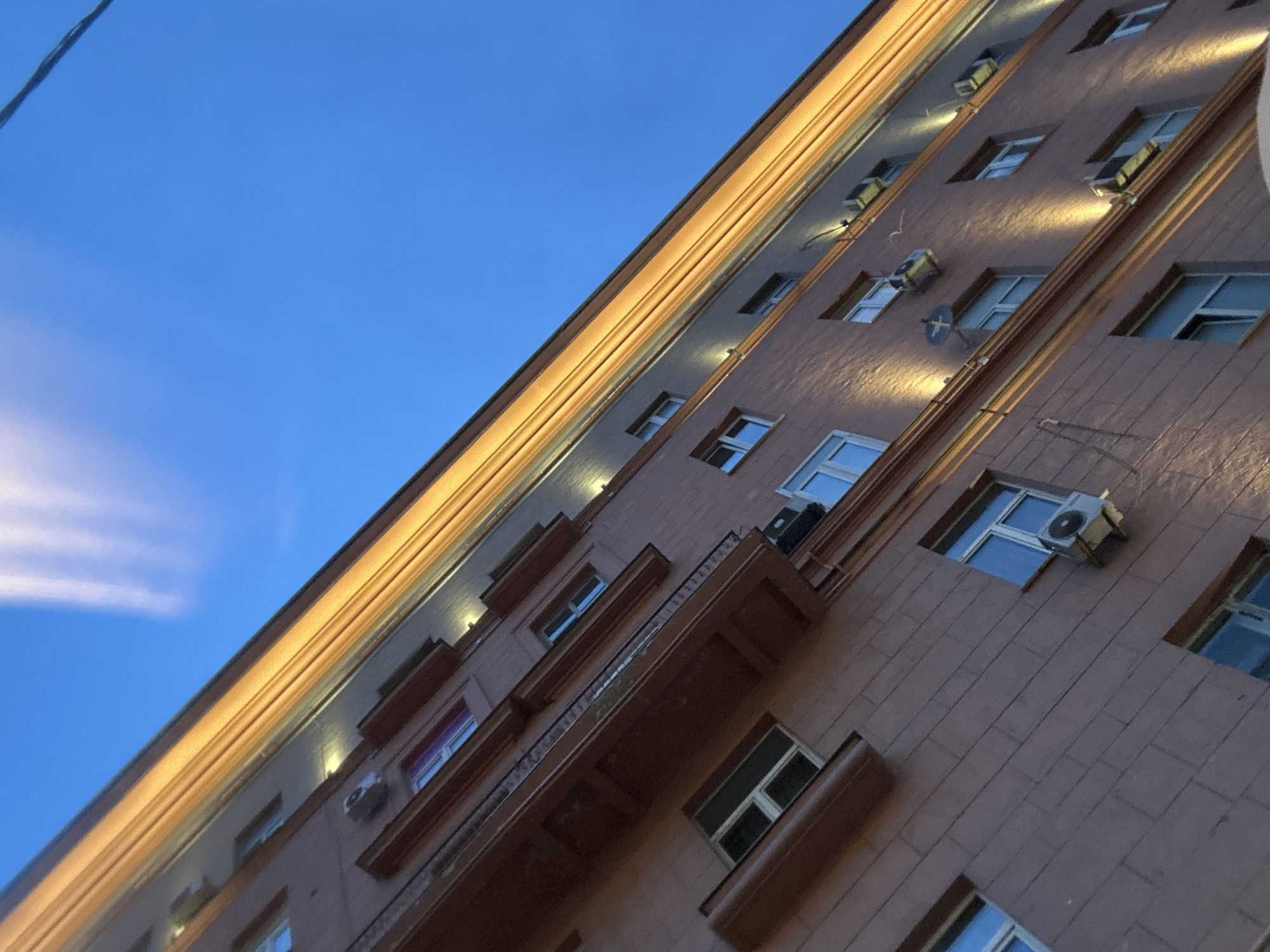 The image depicts the exterior of a building in an urban setting. The building has a multi-story structure with windows, some of which are illuminated, and air conditioning units attached to the facade. There is a decorative ledge or balcony feature present. The building is illuminated by lights, and the overall color palette is muted, with the building appearing to be brownish-red. The sky is visible as a blue gradient, and there is a suggestion of a light cloud formation. The angle of the shot is from below, looking upwards at a tilted perspective. There are power lines near the top left of the image.