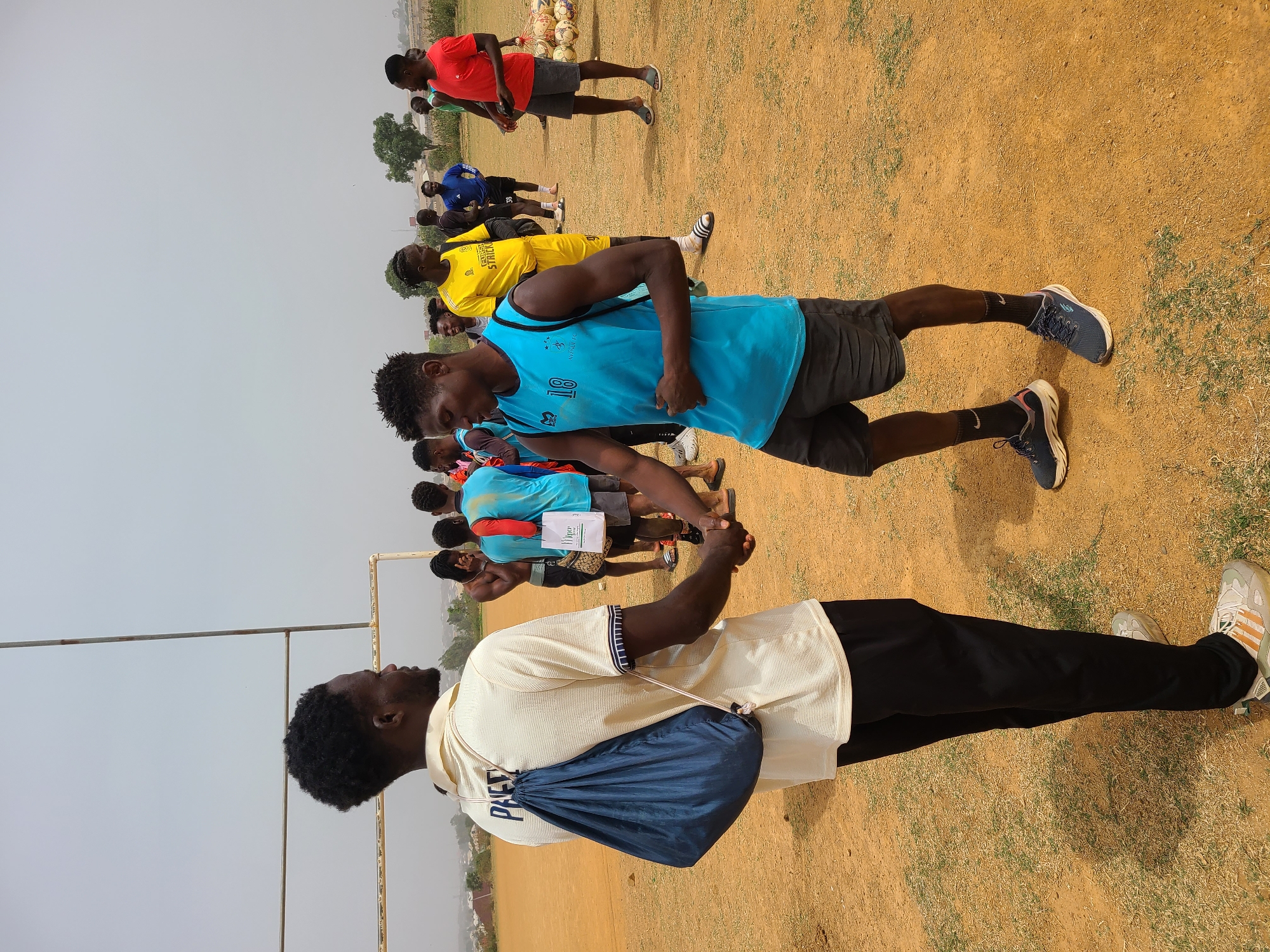A group of young men is gathered on an unpaved, reddish-brown field with sparse patches of dry grass, under a bright, hazy sky. The setting appears to be an outdoor sports ground or training facility. In the background, there are stacks of soccer balls, indicating a soccer-related event. Distant trees and buildings are also visible.
In the foreground, a man in a white or cream polo shirt and black trousers, with a blue drawstring bag bearing the partially visible text "PACE" on his back, is facing another young man in a light blue sleeveless jersey with the number "18" printed on it. They appear to be in conversation or greeting, possibly shaking hands. The man in the blue jersey is wearing black shorts, black Nike socks, and blue athletic shoes.
Behind these two, several other young men are standing in lines or groups. Their attire includes various colored athletic jerseys: yellow (one with partially visible text "STRICK"), red, and other shades of blue, paired with shorts and a mix of athletic shoes, sandals, and slides (one pair featuring the Adidas logo). One participant in the background holds a white paper or board with the visible text "Mengo." The overall impression is of an organized sports activity, likely a training session or tryout, taking place during the daytime in a warm, dry climate.