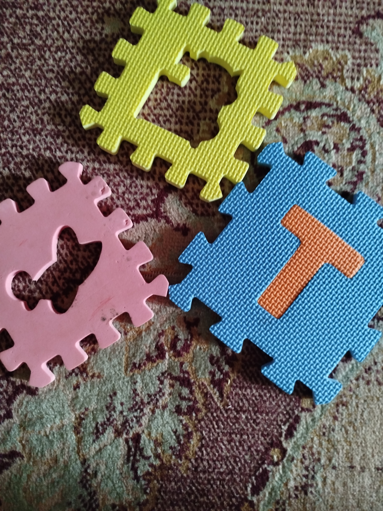 Three puzzle pieces are arranged on a patterned carpet. The puzzle pieces are made of foam and are in different shapes and colors. One piece is yellow and shaped like a square with a heart cutout in the center. Another piece is pink and appears to be a letter "R" with a heart cutout. The third piece is blue and has an orange "T" shape on it. The carpet has a complex pattern with shades of brown, beige, and green. The scene is likely indoors, possibly in a child's play area or a living room. There is no indication of the location being in the city of Крывасельскі сельскі Савет, Belarus.