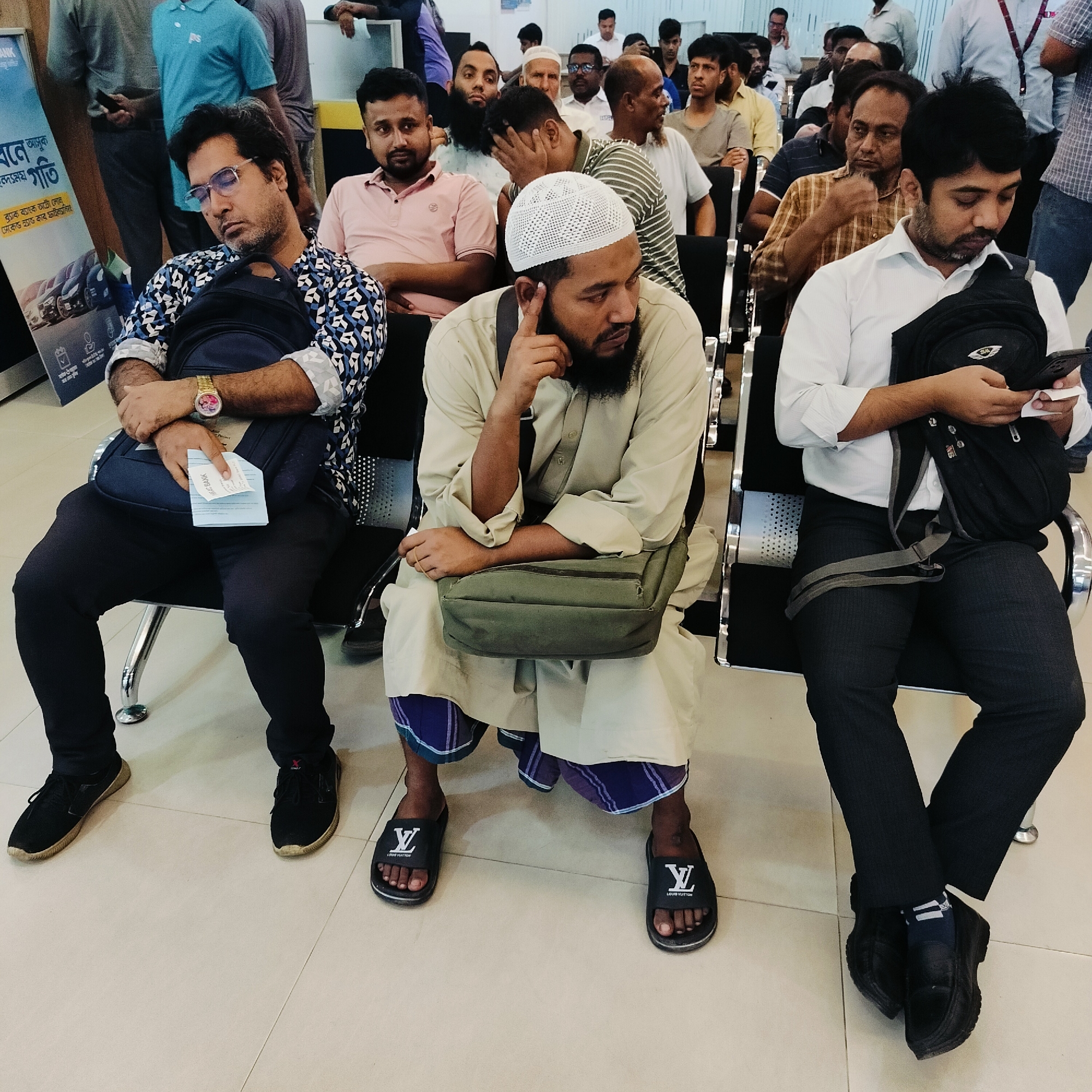The image depicts an indoor waiting area, identified as a bank or financial institution by a partially visible "MAX BANK" advertising banner in the background. Three men are seated in the foreground on black and silver waiting chairs. The man on the left, wearing a blue and white patterned long-sleeved shirt, dark trousers, and black sneakers, has a beard and glasses, and is resting with his head tilted back, holding a dark blue backpack and some papers in his hands. A gold-toned watch is visible on his left wrist. The central man wears a light beige kurta, a purple and white plaid lungi, black "Louis Vuitton" slides, and a white woven prayer cap. He has a beard and is shown in a thoughtful pose, looking to his left, with his right hand near his chin and a green messenger bag across his lap. The man on the right is dressed in a white long-sleeved collared shirt, dark pinstriped trousers, and black dress shoes with blue-striped socks. He has a beard and is observed looking down at a smartphone in his hands while holding a piece of paper, with a black backpack resting on his lap. Numerous other men, primarily of South Asian appearance, are visible in the mid-ground and background, occupying additional seating and standing, indicating a busy public environment. The floor is light-colored tile, and the background features light walls and partitions.