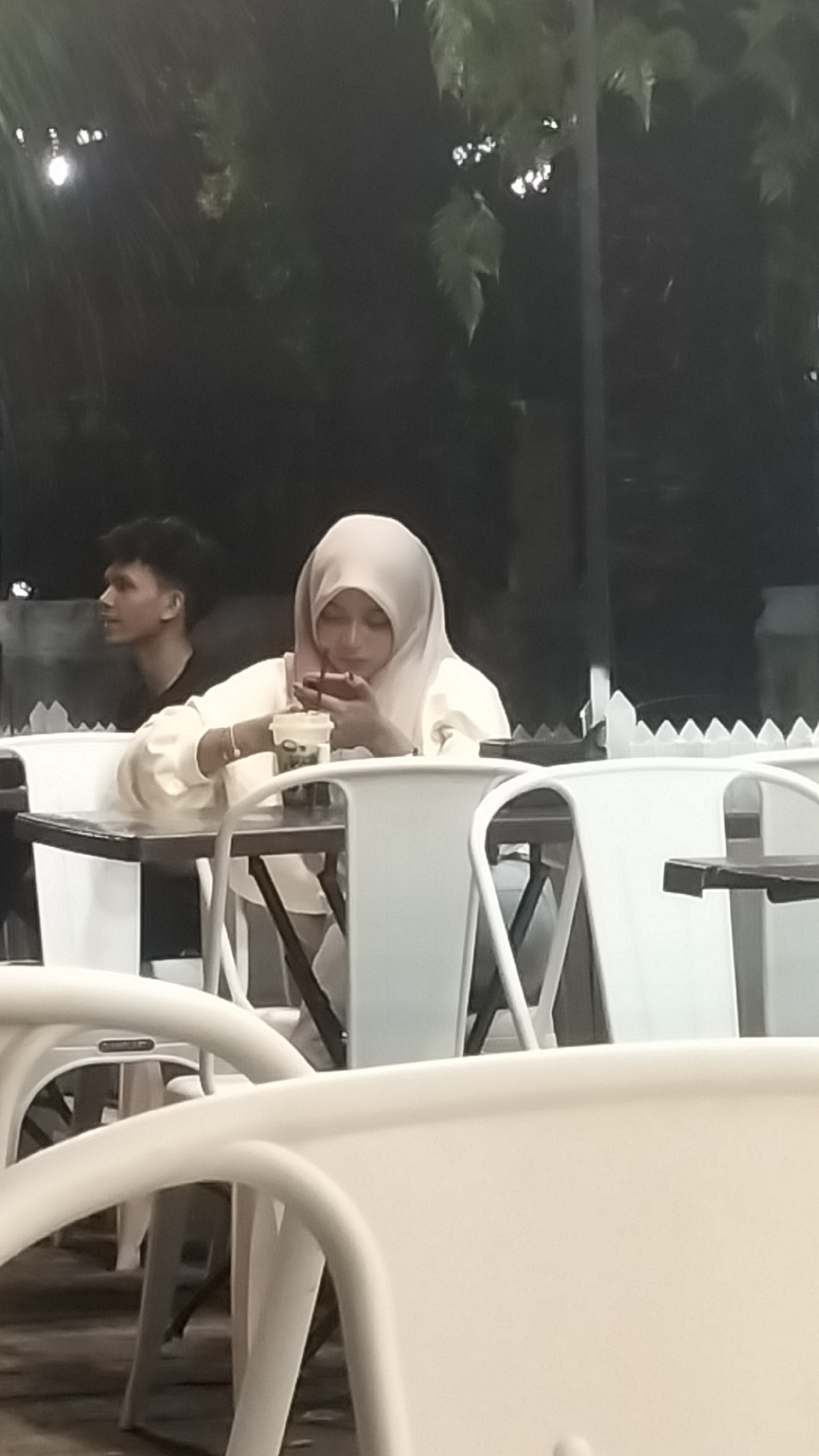 A young woman wearing a light-colored hijab and a white long-sleeved top is seated at a table, looking down at her red smartphone. She is holding a cup with a straw in her right hand, and a bracelet is visible on her left wrist. To her left, a young man with dark hair is looking away from the camera. They are seated at white tables and chairs outdoors, with a blurred background of trees and foliage. The scene appears to be a cafe or restaurant in Lhokseumawe, Indonesia, likely in the evening given the dim lighting.