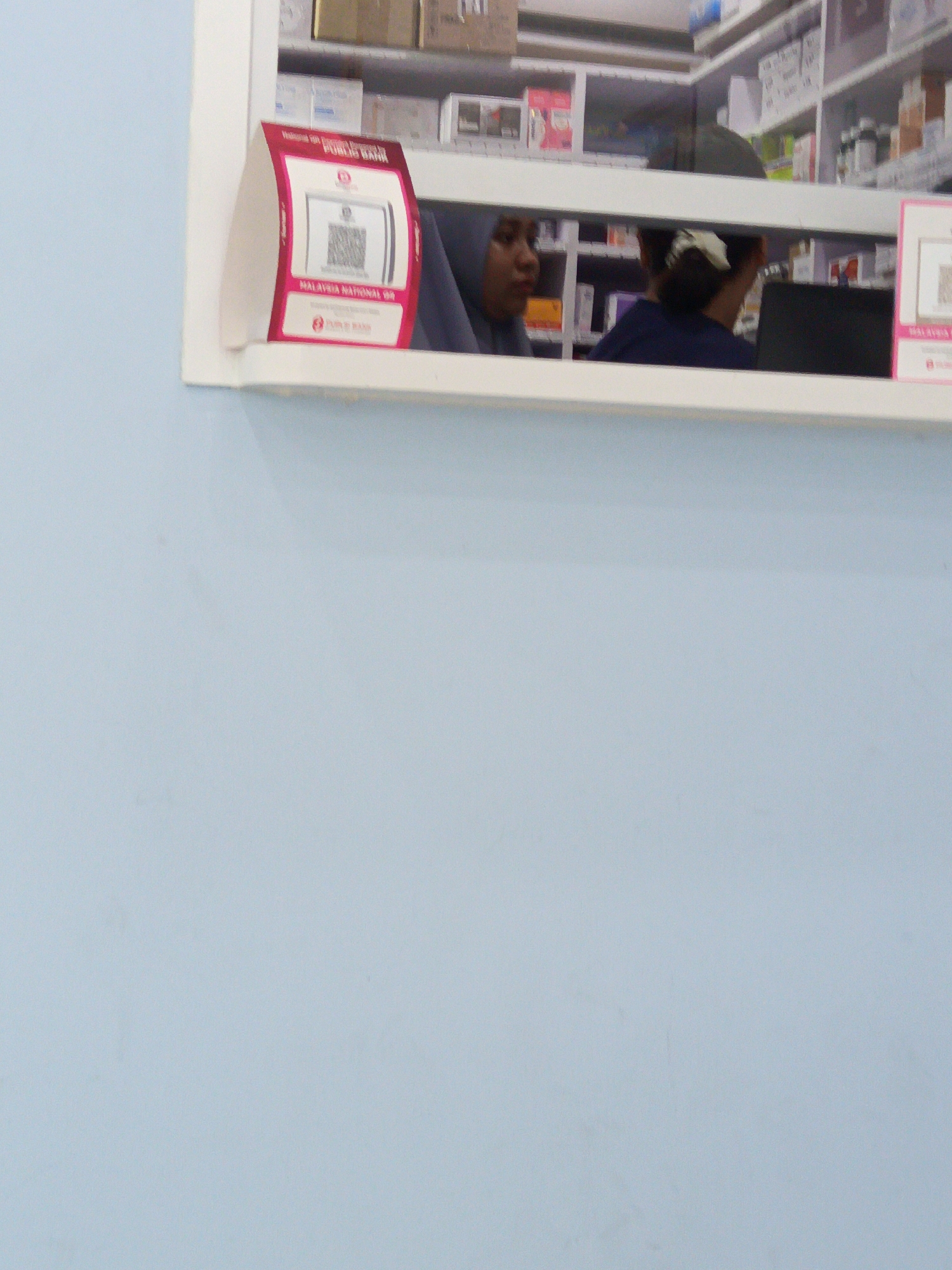 A close-up, eye-level medium shot captures a retail counter with two individuals. In the foreground, a pink and white display sign features a QR code and text that reads "Welcome to Public Bank" and "MALAYSIA NATIONAL QR". To the right of the sign, a woman wearing a blue headscarf is visible, her face turned to the left. Behind her, a person wearing a dark blue shirt and a white scrunchie in their hair is seated and looking down at a laptop. The background reveals shelves stocked with various items, suggesting a retail environment such as a pharmacy or convenience store. The overall lighting is bright, indicating an indoor setting. The location is identified as a Public Bank branch in Kuala Lumpur, Malaysia, based on the signage.