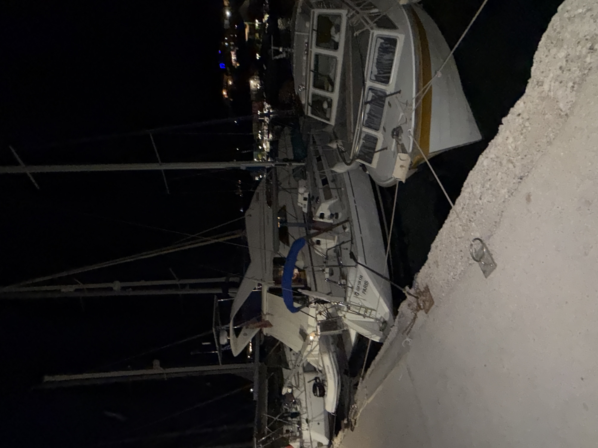 The image depicts a nighttime scene at a marina in the Municipal Unit of Palairos, Greece. Several boats are docked along a concrete quay. In the foreground, a white motorboat with a yellow stripe on its hull is moored. Behind it, a white sailboat named "PARIS" is visible, with a blue canopy over its cockpit. Further back, another sailboat and a small inflatable boat are also docked. The background shows a dimly lit town with lights from buildings and streetlights, suggesting a residential or commercial area near the harbor. The overall atmosphere is calm and still, with the boats reflecting faintly on the dark water.