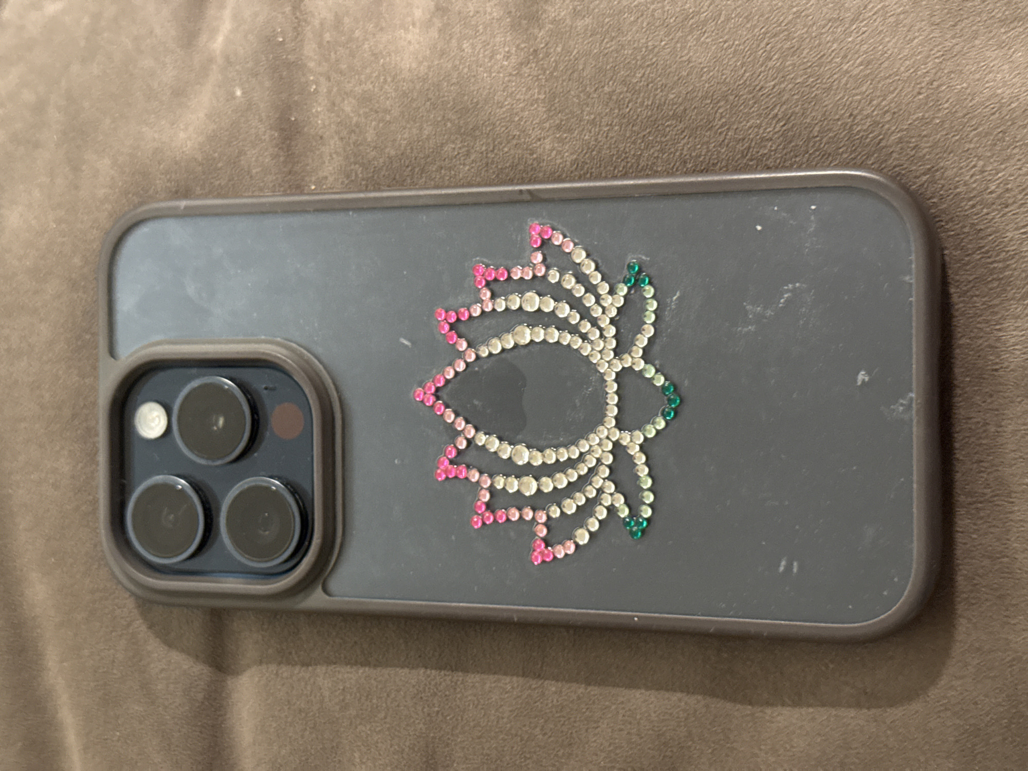 A close-up shot shows the back of a dark grey iPhone with a lotus flower made of rhinestones. The lotus flower has pink rhinestones on the top petals, white rhinestones in the middle, and green rhinestones on the bottom petals. The phone is lying on a brown textured surface, likely a blanket or rug. The phone's camera module, located in the upper left corner of the image, features three circular lenses and a small circular sensor. The background is out of focus. The image does not depict any elements related to the city of Бараўлянскі сельскі Савет, Belarus.