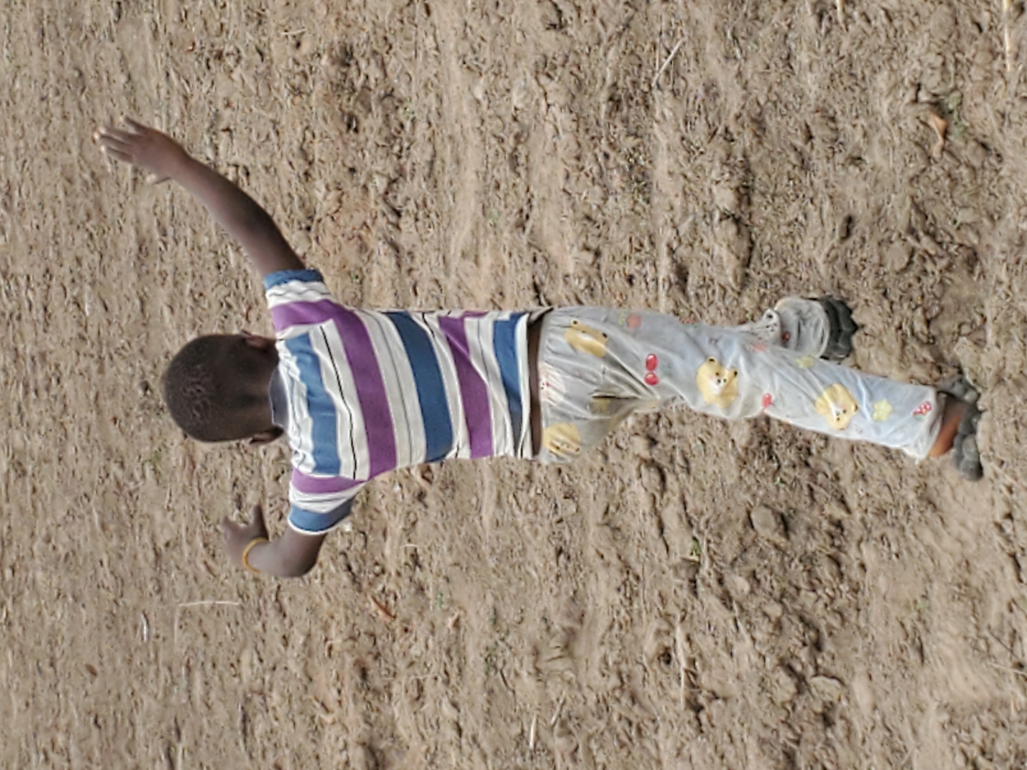A top-down view captures a child lying on dry, uneven, earthy ground in Mararaba, Nigeria. The child, who has dark skin and short dark hair, is positioned on their back or side with arms and legs outstretched. Their right arm is extended upwards, while the left arm is bent at the elbow, with the hand resting near their head. This pose suggests a moment of play or rest outdoors.
The child is dressed in a short-sleeved, striped shirt featuring vertical bands of blue, white, and purple or maroon. They wear light-colored pants adorned with patterns that include yellow bear faces or similar animal motifs, alongside red circular or oval shapes. Dark-colored footwear, possibly sandals or simple shoes, is visible on both feet. A thin yellow bracelet or band is worn on the child's right wrist.
The ground beneath the child is a brownish-tan color, typical of dry soil, with visible small stones, clumps of earth, and sparse dried plant matter. The overhead perspective provides a clear view of the child and their immediate surroundings. The lighting indicates it is daytime, and the dry nature of the ground suggests warm or arid conditions. No visible text is present in the image.