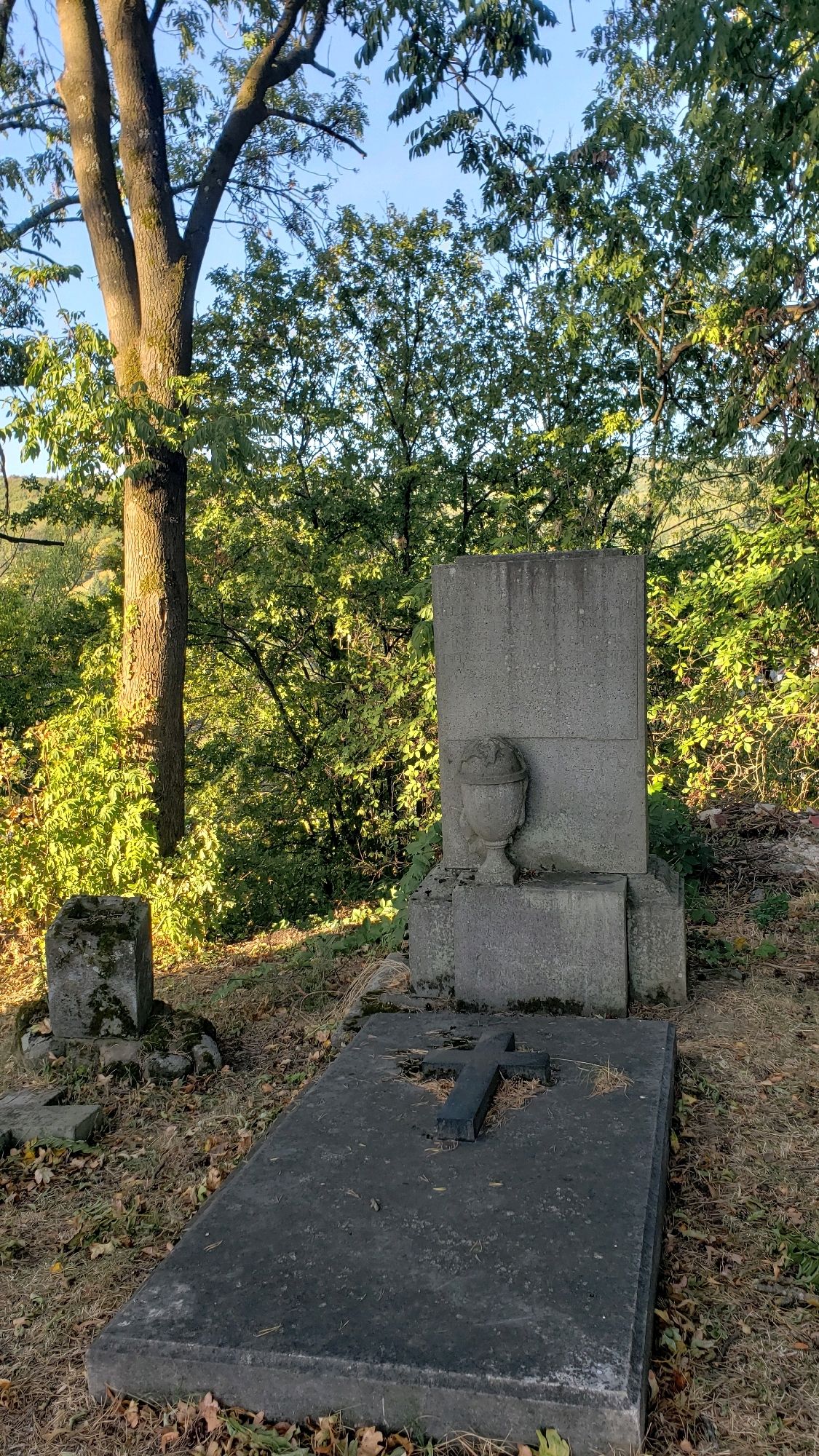 The image depicts a tombstone and grave in a cemetery. The tombstone is made of grey stone and has a rectangular base with a larger rectangular slab on top. A small urn-shaped decorative element is placed on the front of the base, near the top. In front of the tombstone, a flat, rectangular stone slab covers the grave. A dark, cross-shaped object is placed diagonally on this slab. The ground around the grave is covered with dry leaves and grass. In the background, trees with green foliage are visible, suggesting an outdoor setting, likely a cemetery. There are no people visible in the image. The scene appears to be in a natural, somewhat overgrown environment.