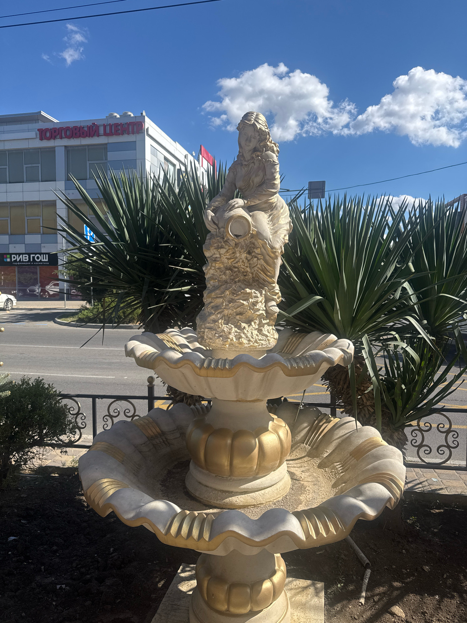 A tiered, ornate fountain with a statue of a woman holding a jug stands in the foreground. The fountain is cream-colored with gold accents and appears to be made of stone or concrete. Behind the fountain, two large, spiky palm trees flank the statue. In the background, a modern, white building with large windows is visible. Red lettering on the building reads "ТОРГОВЫЙ ЦЕНТР" (Shopping Center). To the left of the building, a sign for "РИВ ГОШ" (a Russian cosmetics and perfume retailer) is partially visible. A road with a white car is visible on the left side of the image, and a black metal fence with decorative scrolls runs along the front of the fountain. The sky is blue with scattered white clouds. This scene is located on a sunny day in the city of Anapa, Russia.