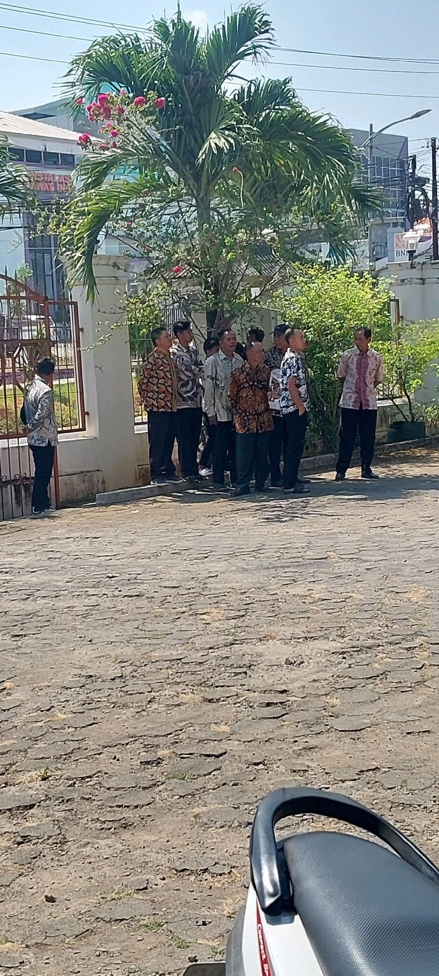 In Keling, Indonesia, on a sunny day, a group of approximately nine men, all dressed in batik shirts and dark pants, are standing in a line outdoors. They appear to be waiting or observing something to their right. The ground is paved with textured, interlocking stones. Behind the men, a white wall with a metal gate stands, and beyond that, a large palm tree with pink flowers is prominent. In the background, a modern building with glass windows and signage is visible, along with utility poles and electrical wires. A portion of a scooter's seat and handlebars is in the foreground, on the right side of the frame.