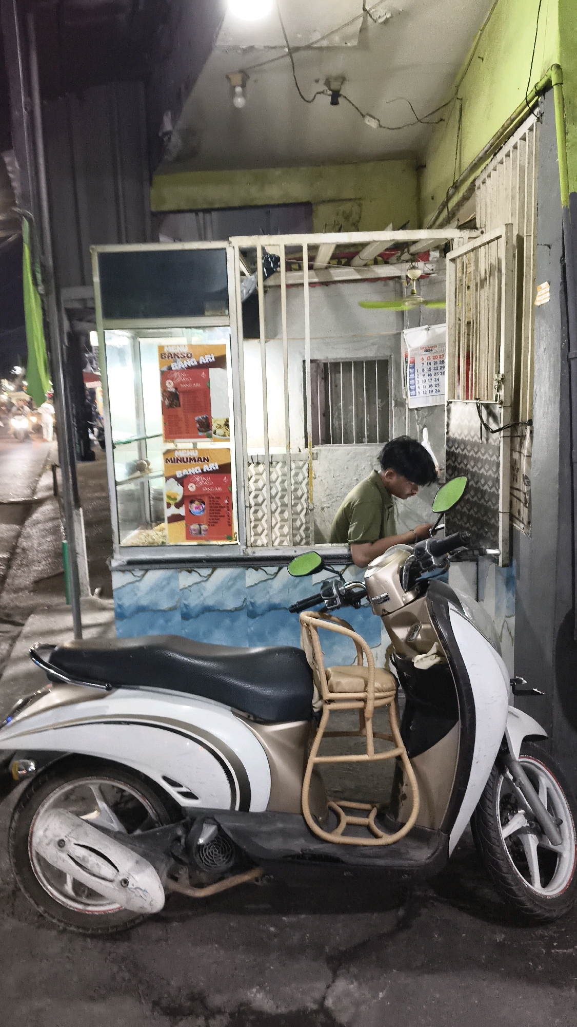 In Bekasi, Indonesia, a scooter is parked in front of a small food stall. The stall has a glass front displaying menus for "BAKSO BANG ARI" (Beefball Bang Ari) and "MINUMAN BANG ARI" (Drink Bang Ari). A young man wearing a green t-shirt is leaning out of the stall's window, looking down at his phone. The stall is attached to a building with a green facade and a metal grill. A calendar is visible inside the stall. The scooter has a white and gold body and a small child's seat made of wicker attached to its front. The ground is asphalt, and the scene appears to be at night, with artificial lighting illuminating the area.