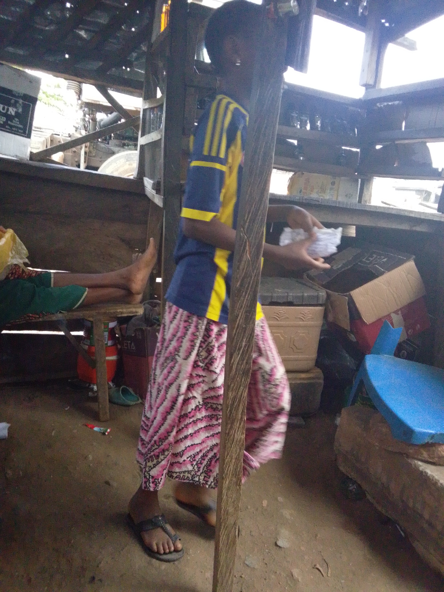 The image depicts an indoor scene within a rudimentary wooden structure, likely in Lokoja, Nigeria, open to the bright daylight outside. The floor is dirt.
In the foreground, a person stands, facing slightly away from the camera. They wear a blue and yellow striped short-sleeved shirt, pink and white patterned trousers or a long skirt, and dark flip-flops. Their hands hold a bundle of white papers or fabric, positioned above some stored items. Their face is partially visible, with dark skin and hair, looking towards the right side of the frame.
To the left, a second person is resting, lying on their back with their bare feet propped up on a simple wooden stool or low table. They are wearing green shorts.
The immediate surroundings are cluttered with various objects. Underneath the resting person's feet, a red bucket or container is visible, and next to it, a light blue pair of flip-flops. Near the standing person, a beige ceramic pot with decorative patterns sits atop something, and next to it, an open cardboard box containing other items. Further right, a bright blue plastic stool is on the ground.
Through gaps in the wooden structure, possibly a wall or roof, bright daylight streams in. Parts of a vehicle, including tires and machinery, are visible outside, suggesting an outdoor area immediately adjacent to the structure.
Visible text includes "UN" on a white box or sign in the upper left, and "ETA" on the red container near the resting person's feet. The scene appears to be an everyday moment in a casual, possibly commercial or residential, setting during the daytime.