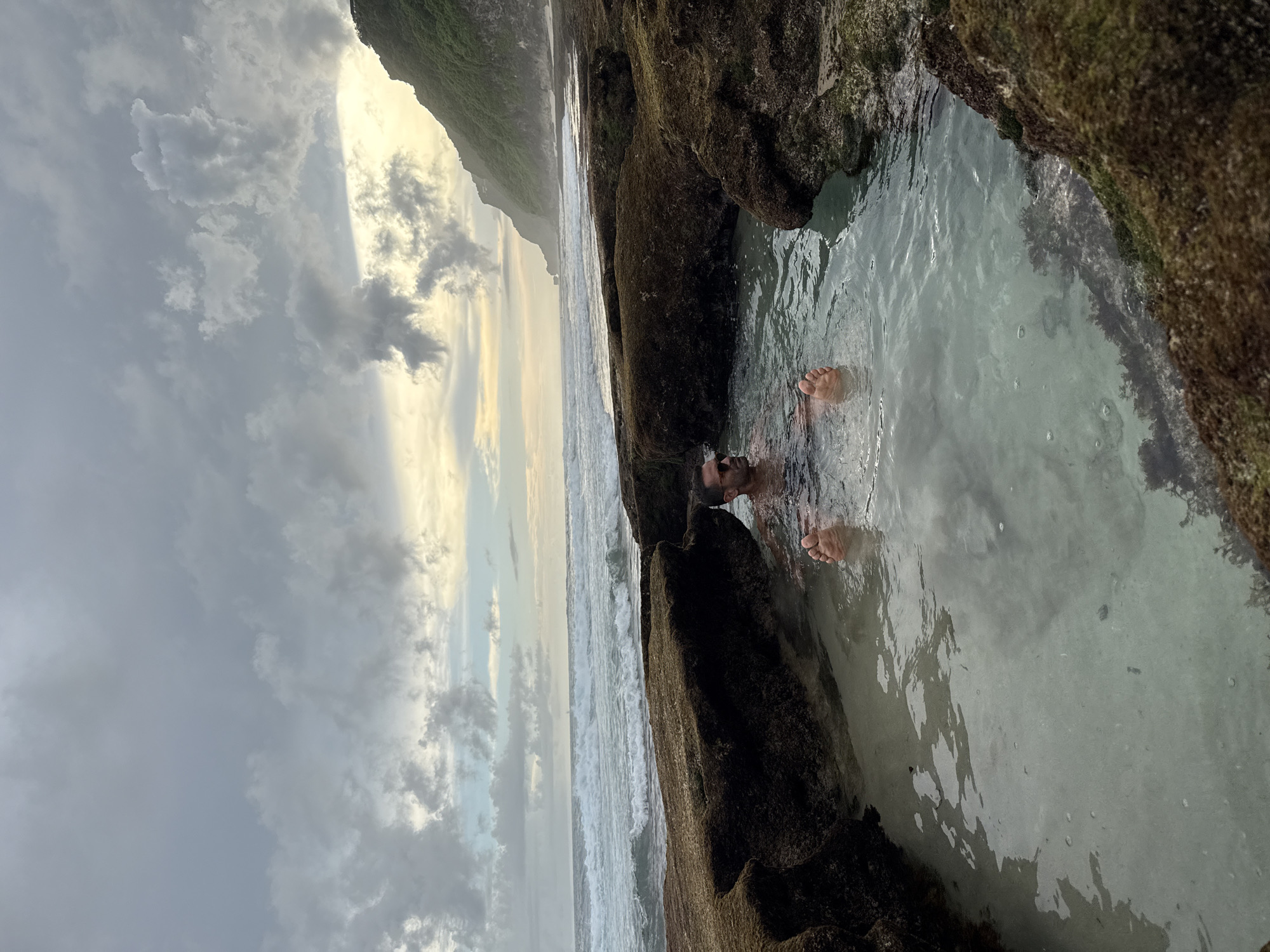 A person is immersed in a tide pool at the edge of the ocean near Pecatu, Indonesia. Rocky, moss-covered formations define the pool’s edges. The person's feet and upper body are visible above the surface of the water. The ocean extends to the horizon under a sky with cloudy conditions. The coastline is on the left.