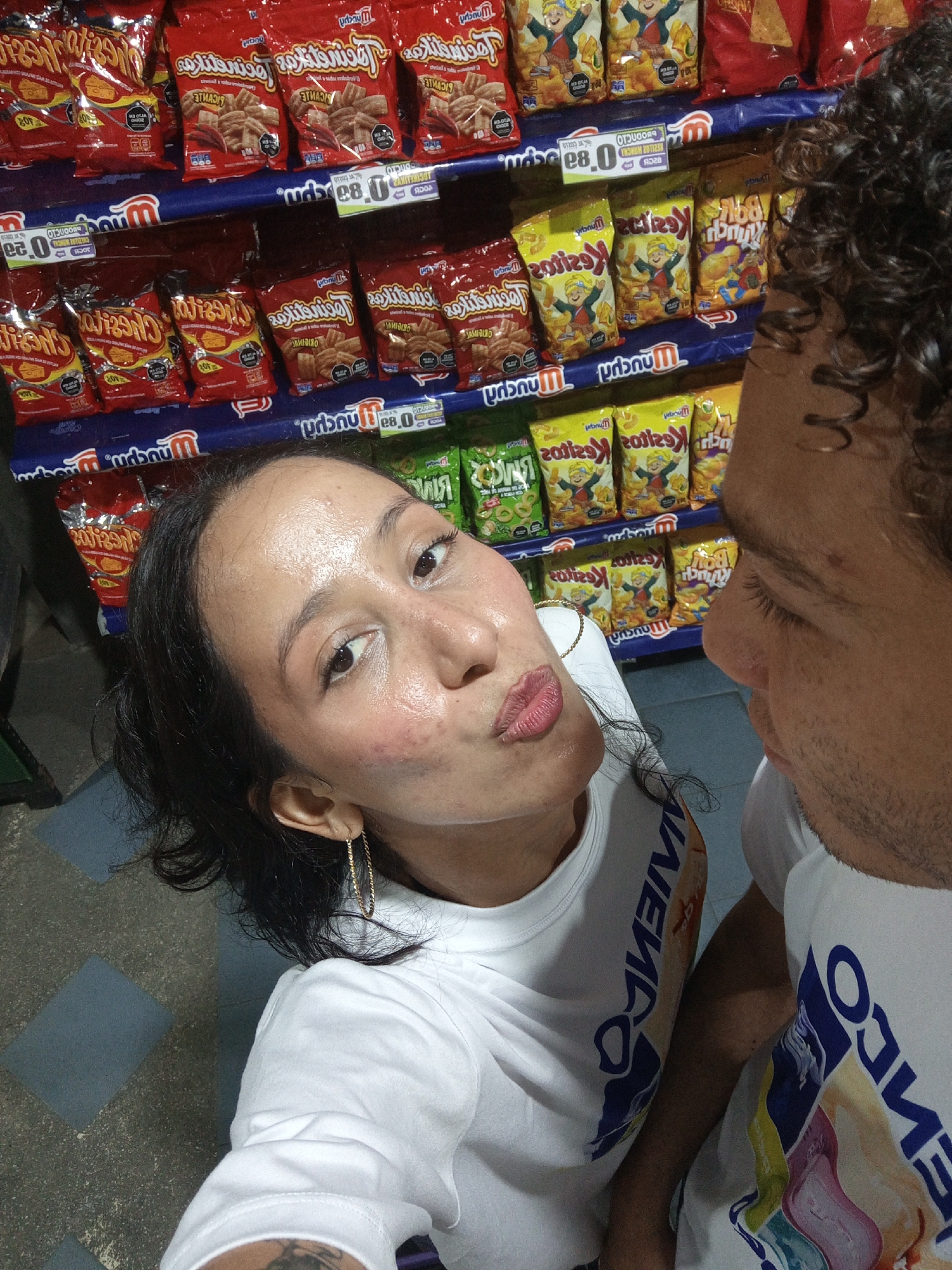 This photograph appears to be a selfie taken indoors, possibly in a small grocery store or a convenience store. The focus is on a couple, with the woman in the foreground making a kissing face towards the camera. She has dark hair, fair skin, and is wearing a white t-shirt and gold hoop earrings. The man is partially visible on the right side of the frame, with curly dark hair and wearing a matching white t-shirt. The woman has some blemishes on her face. Behind them is a shelf stocked with various snack items, predominantly bags of chips. The shelves are labeled with prices. The floor appears to be tiled. The brand name “Munchy’s” is prominently displayed on the snack packaging. The text on the shirts appears to be mirrored, spelling "CONVENIO". The location, Porlamar, Venezuela, is derived from this brand, “Convenio".