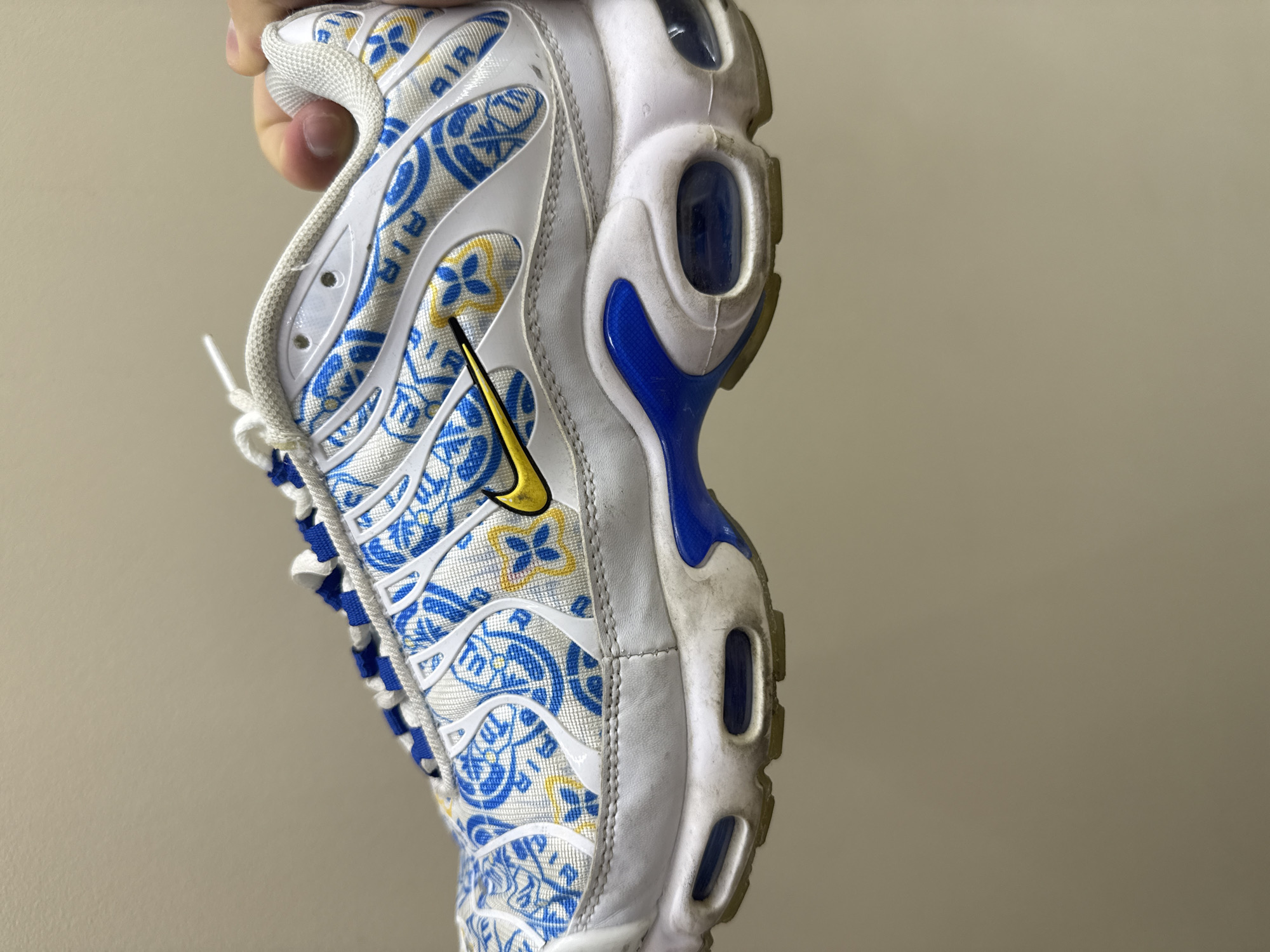 A person's hand is holding a Nike Air Max Plus sneaker. The sneaker has a white base with a blue and yellow tiled pattern. The Nike swoosh is yellow. The shoe is slightly dirty, particularly around the sole. The background is a plain, light-colored wall. There is no indication of location in Burgas, Bulgaria.