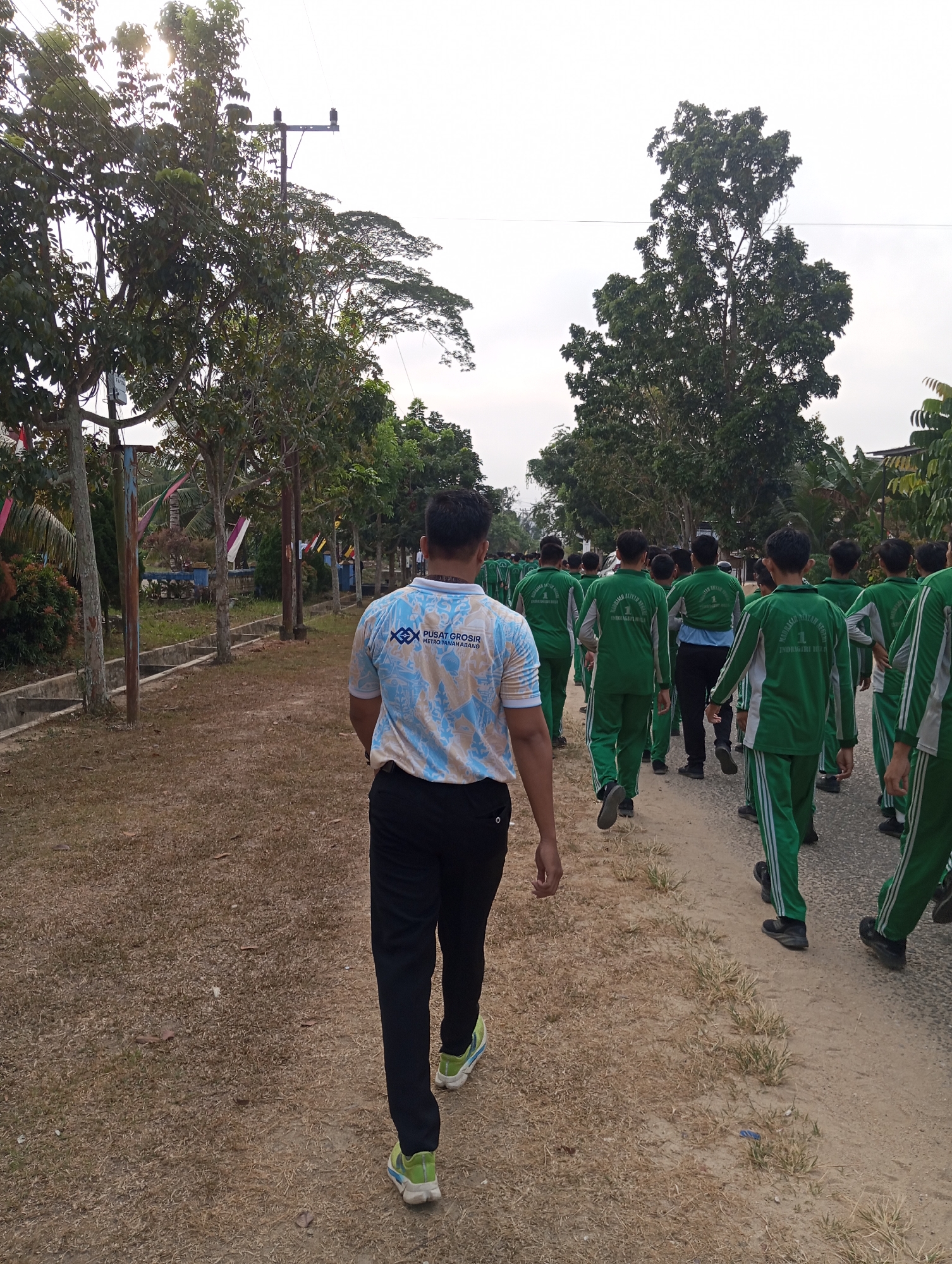 A group of young males, likely students, are walking in a line on a dirt path. They are all wearing matching green tracksuits with white stripes. The person in the foreground is wearing a light-colored t-shirt with a blue pattern and black pants. The environment appears to be a rural area with trees lining the path. There are also utility poles and wires visible. The sky is overcast. The group is walking away from the camera. The scene is likely set in Pematang Reba, Indonesia, given the presence of the utility poles and the vegetation.