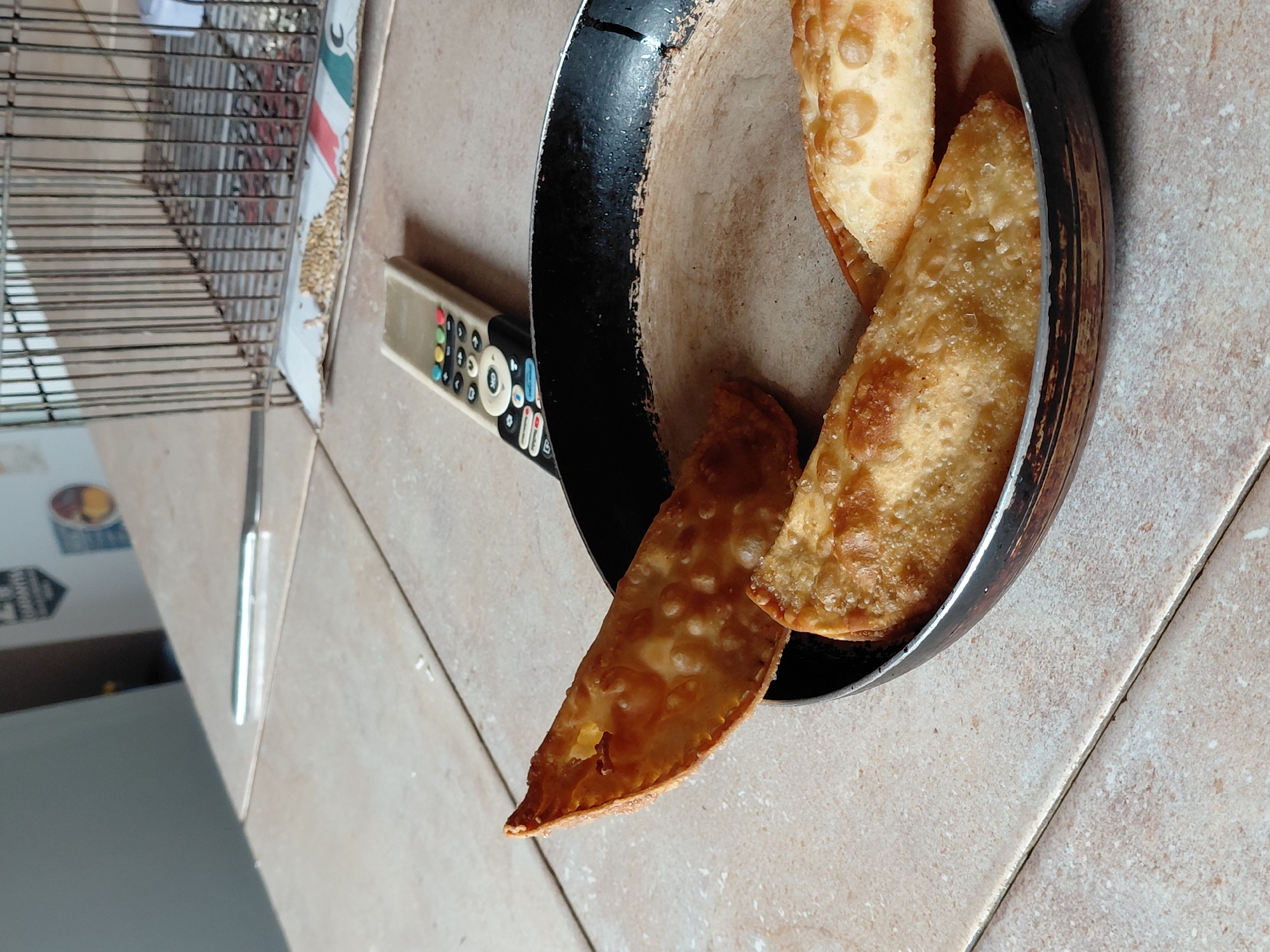 The image captures a casual indoor scene, likely in a home in Santa Ana, Venezuela, taken from a slightly elevated and tilted perspective. The primary focus is a well-used black frying pan containing three golden-brown, fried pastries, resembling empanadas or pasteles. These pastries exhibit a bubbly, crispy texture, suggesting they are freshly prepared. One of the pastries has a small opening, revealing a hint of its filling.
To the left of the pan, a beige remote control rests on the tiled floor. Visible text on the remote includes "SAMSUNG" and dedicated buttons for streaming services like "NETFLIX" and "Prime Video," along with a "WWW." button and other common remote functions.
Further to the left, a metal bird cage is partially visible, indicating the presence of a pet bird. Below the cage, a scattering of bird seeds is spread on what appears to be a piece of cardboard or a tray. The floor throughout the scene is covered with light-colored, possibly beige or cream, square tiles. The lighting suggests it is daytime. No people are present, and the scene is a still life, portraying everyday domestic items.