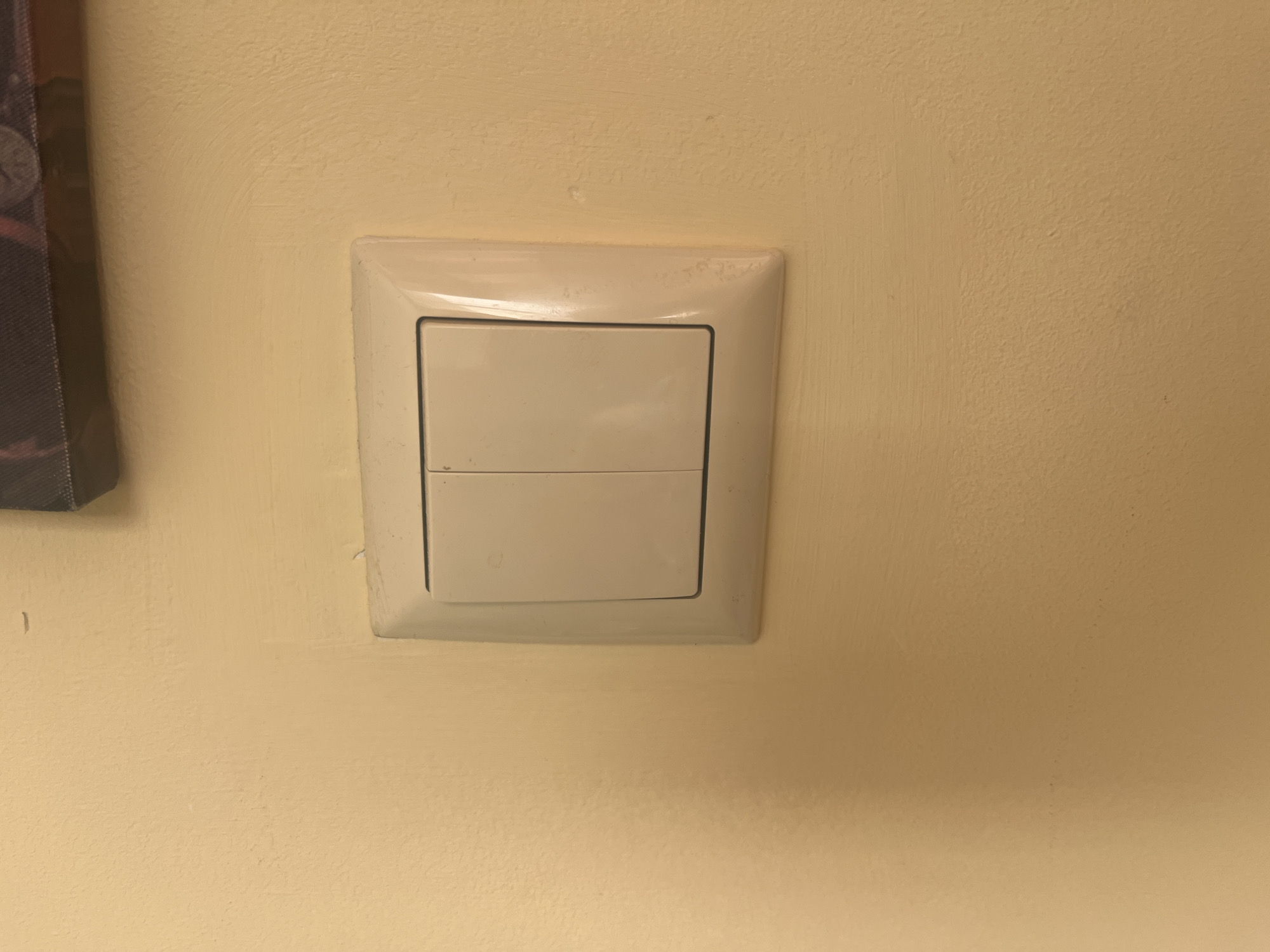 A white plastic double light switch is mounted on a textured, light yellow-beige painted wall in Burgas, Bulgaria. The switch faceplate and two rectangular rocker buttons exhibit signs of wear, including localized discoloration and grime accumulation around the edges. The wall's paint finish is uneven, displaying visible brush or roller marks and minor surface imperfections. A dark, framed object with a textured surface and subtle circular patterns is partially visible in the upper right portion of the image, positioned above the light switch.