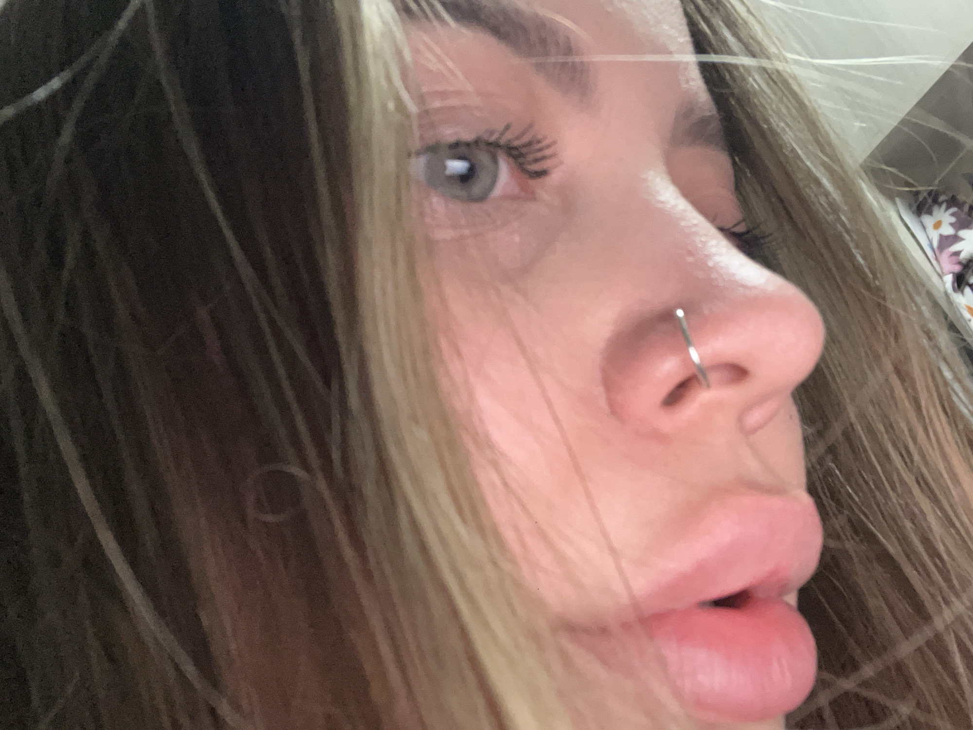 A close-up, angled portrait features the left side profile of a person's face, focusing on the eye, nose, and lips. Long, multi-toned hair, appearing to be light brown with blonde highlights, frames the face, suggesting movement. The person's left eye has a light-colored iris, possibly grey or light blue, with noticeable eyelashes, likely enhanced by mascara, looking subtly upwards. A simple, metallic silver hoop nose ring is worn in the left nostril. The lips are full, pink, and slightly parted. The skin tone is fair, and the overall expression is neutral and calm. The background is out of focus, with a faint floral pattern visible in the upper right, suggesting an indoor setting with patterned fabric or wallpaper. No specific activity is depicted beyond the pose for the photo. There is no visible text within the image. The location context provided is Novopokrovskaya, Russia.