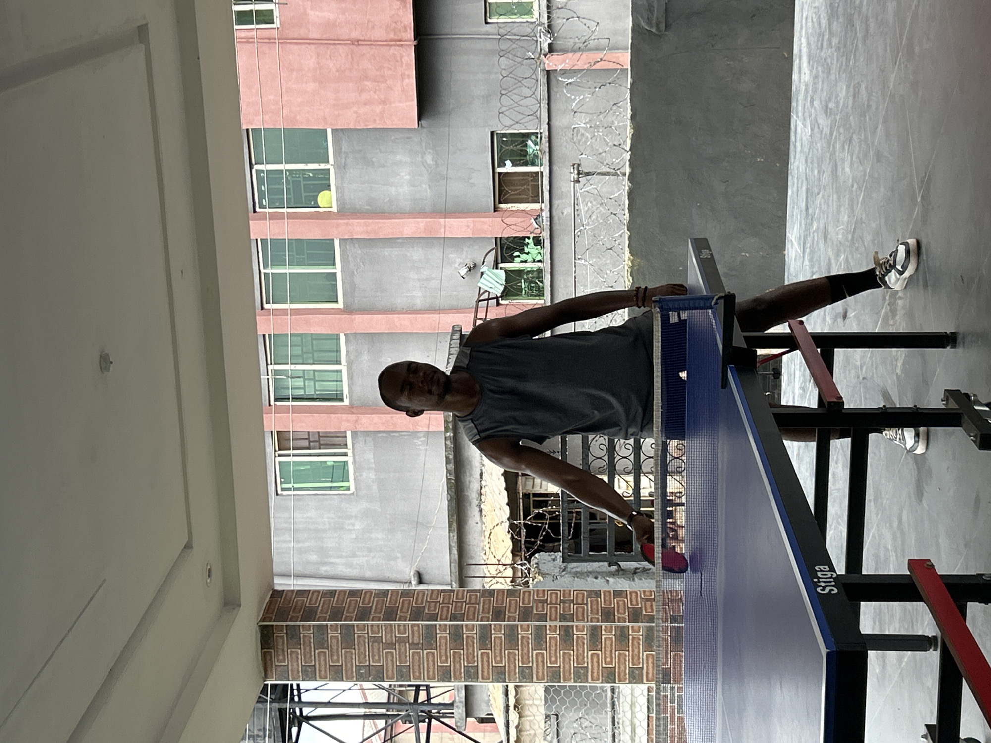 A dark-skinned man stands on a tiled floor beneath an overhang, playing table tennis. He is wearing a grey sleeveless shirt, black socks, and white and black athletic shoes. He holds a red table tennis paddle in his right hand, poised to strike a ball over the net of a blue Stiga table. Behind the table, a grey concrete wall is visible, topped with barbed wire. Further back, a building with pink and grey facades features several windows. The scene appears to be in an outdoor recreational area.