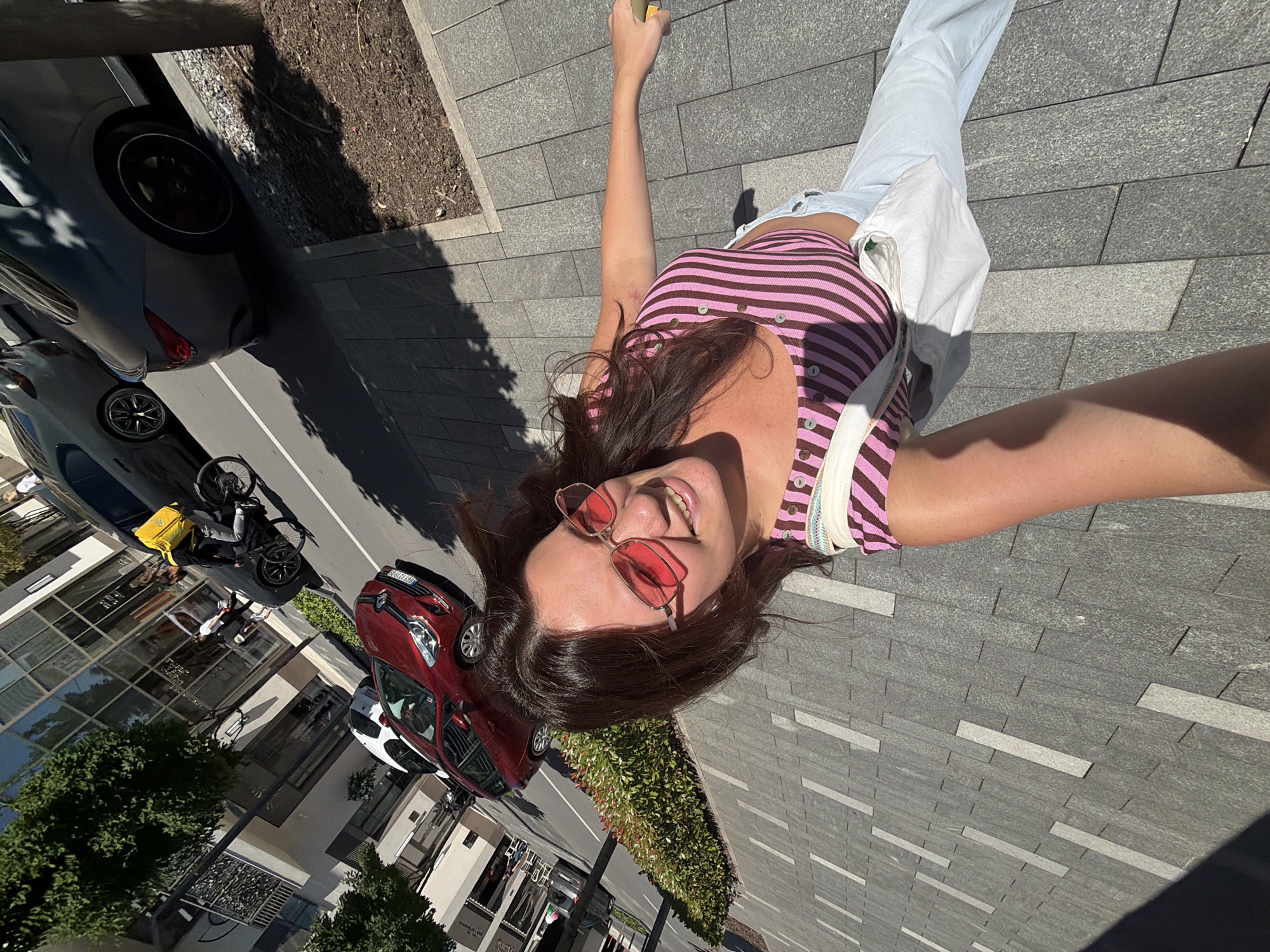 A young woman with long, dark brown hair and pink-tinted sunglasses is taking a selfie outdoors on a paved city sidewalk. She is wearing a pink and brown striped tank top with white pants and a white bag slung over her shoulder. Her right arm is extended, holding what appears to be a phone. In the background, a red car is parked on the street, and further down the street, a person on a bicycle with a yellow delivery bag is visible. The environment suggests a sunny day in an urban setting, with buildings and trees lining the street. The location is identified as Belgrade, Serbia.