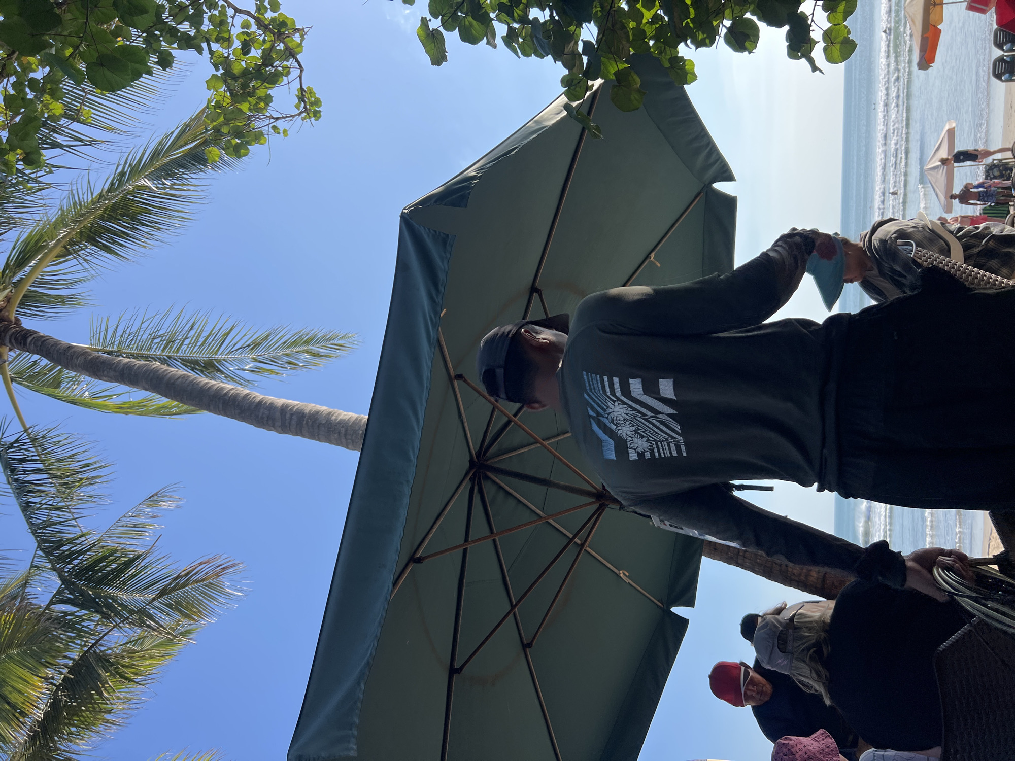 The image shows a beach scene in Kuta, Indonesia. In the foreground, a person wearing a dark long-sleeved shirt and a baseball cap stands under a large green umbrella, facing away from the camera. The umbrella's structure is visible. Behind this person, other individuals are partially visible, some wearing hats. The background reveals the ocean with gentle waves and a clear blue sky. Palm trees and foliage frame the scene, suggesting a tropical environment. Several beach umbrellas and lounge chairs are scattered on the sand, indicating a public beach area. The overall atmosphere is relaxed and sunny.