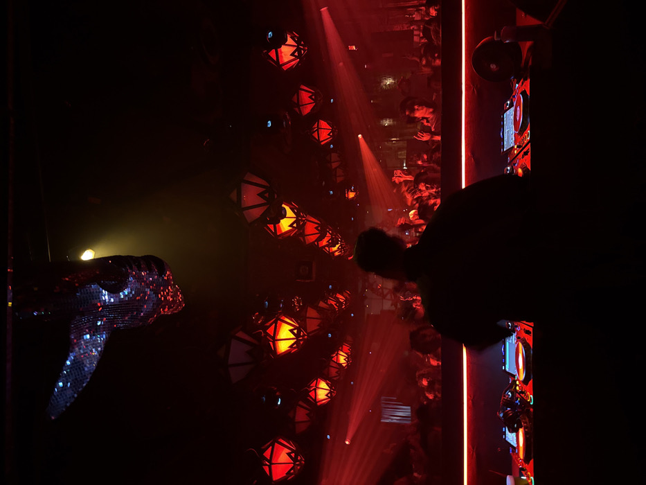 This image captures the interior of a nightclub or lounge, identified as likely "Kiss Kiss Bang Bang" within the LINE LA Hotel in Koreatown, Los Angeles, United States of America.
The scene is dominated by a dark, hazy environment illuminated primarily by intense red light. In the foreground, a male individual, presented as a silhouette, stands with his back to the camera, positioned behind a DJ console. The console is equipped with two illuminated turntables/CDJs and a central mixer, indicating active performance. A beverage cup is visible on the left side of the console.
Above and behind the DJ, a dense crowd of approximately 50-70 individuals is visible, mostly as silhouettes or dimly lit figures, engaged in activity consistent with dancing, with several hands raised in the air. This crowd occupies the midground, separated from the DJ by a horizontal red LED strip outlining a stage or platform. Red laser or spotlight beams cut through the ambient haze above the crowd.
The ceiling features numerous geometric lanterns, predominantly red with some warm yellow interiors, suspended overhead. In the upper left quadrant, an elongated disco ball reflects fragmented light. A single, focused beam of warm yellow light emanates from a fixture near the disco ball. The overall atmosphere suggests a high-energy entertainment venue.