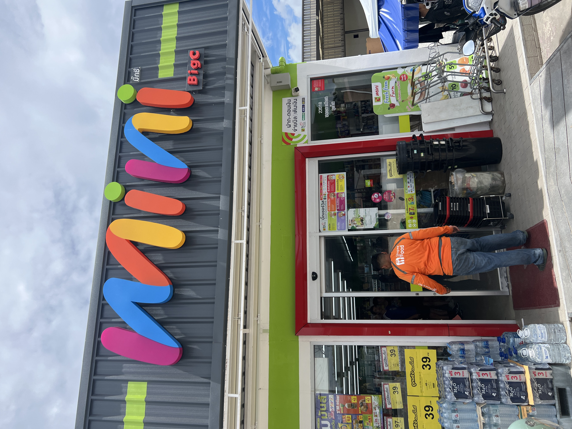 A Mini Big C convenience store is shown in Chalong, Thailand, under a cloudy sky. The store has a prominent, multi-colored "mini" logo on its facade, with "Big C" written in red text below it. A Shopee Food delivery person wearing an orange jacket and blue jeans is standing by the entrance, facing the store. Inside the store, shelves stocked with goods are visible. In front of the store, stacks of bottled water are displayed, along with shopping carts and black trash cans. A blue canopy is set up to the right of the entrance, partially obscuring a motorcycle.