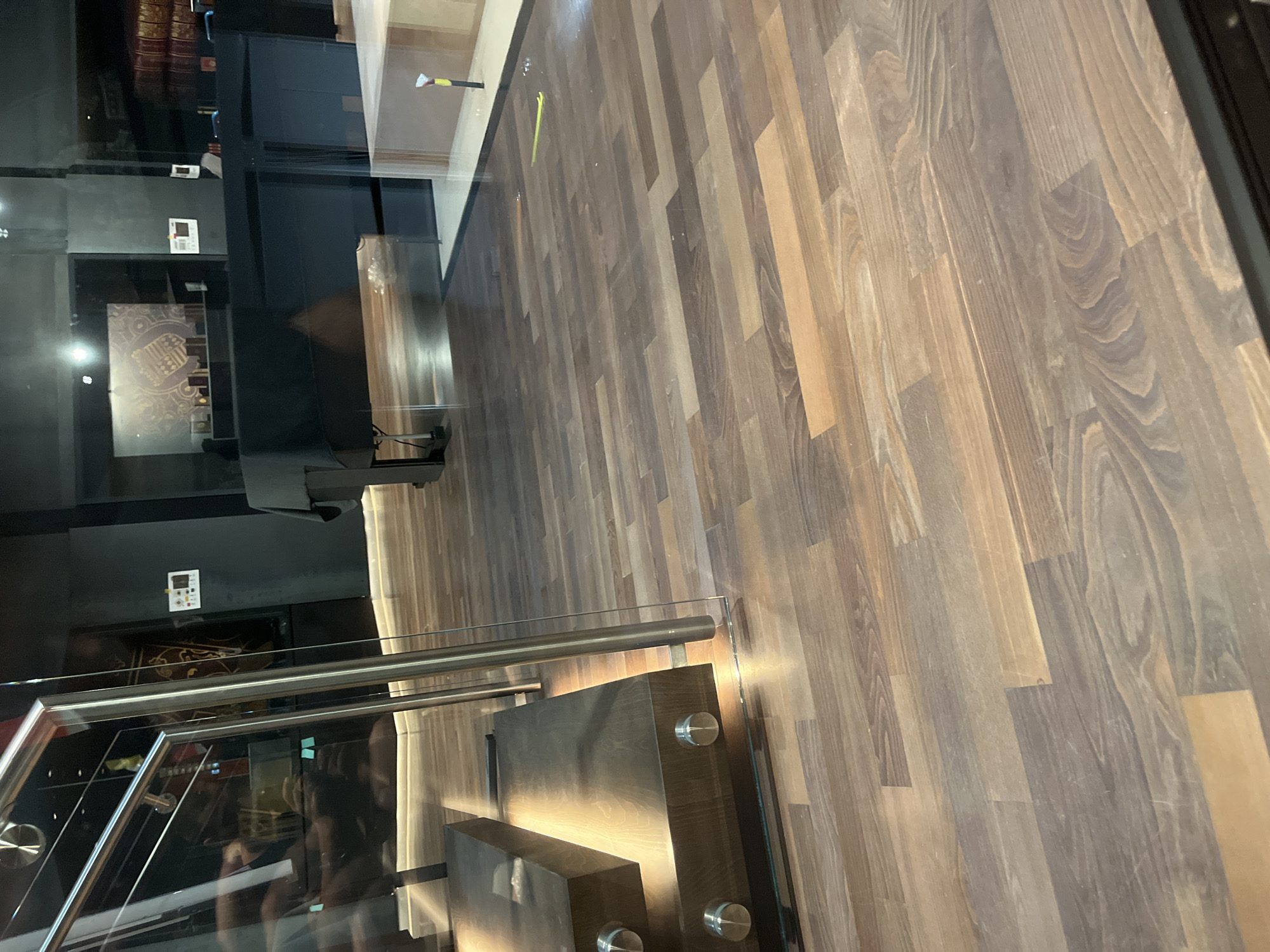 This is a tilted, low-angle, eye-level, full shot of a staircase and a wooden floor in what appears to be a modern building. The staircase has glass sides and a metallic handrail. The steps are made of dark wood and are illuminated from underneath, casting a warm glow. The floor is made of long, narrow wooden planks with a natural grain pattern, varying in shades of brown and grey.
In the background, there are reflections in the glass. One reflection shows what looks like a wall with some framed items and a display cabinet. Further in the background, books are visible on shelves. Another reflection shows a person taking the photo, with dark hair and wearing dark clothing, holding a phone.
There is a yellow object on the floor near the top edge of the image, and a small white electronic device with a few buttons on a dark surface in the background, possibly a control panel.
The lighting is artificial, with the illuminated staircase and the overall tone suggesting it could be evening or indoors. The location cues are minimal, but the scene appears to be an interior space, possibly a museum, library, or upscale commercial building. No specific activity or event is clearly depicted beyond the implied presence of people navigating the space. The mood is neutral and clean, with an emphasis on the modern design and materials.