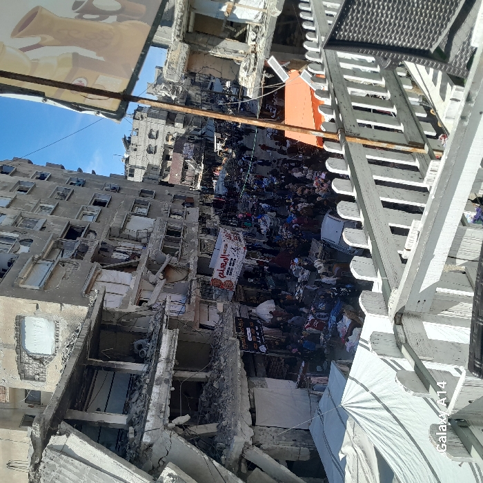 This image provides an elevated view of a busy street in Khan Yunis, Palestinian Territory, flanked by heavily damaged multi-story concrete buildings. The buildings on both sides exhibit significant structural compromise, with large sections of walls collapsed, exposed rebar, missing facade elements, and numerous broken windows, indicating widespread destruction. Rubble and debris are evident around their bases and within the damaged interiors.
The street below is densely populated with a large crowd of individuals, primarily adults, engaging in commerce within a bustling open-air market. Numerous temporary market stalls are set up, displaying a variety of goods, including textiles and household items, arranged on tables and tarps. People are observed walking, standing, and interacting throughout the market area. A light-colored sedan is partially visible amongst the crowd on the street.
A large white banner displaying red Arabic text, "المعلم" (Al-Mu'allim), is strung across the street overhead. Additional smaller signs with Arabic script are visible on storefronts. In the upper right portion of the frame, a partially obscured large sign features an illustration of traditional pottery. An orange awning is visible on the right side of the street at a lower level, with small solar panels mounted nearby. In the immediate foreground, a white picket-style railing frames the bottom and left, indicating the elevated vantage point. A large white tarp or tent structure is present in the lower-left foreground. The sky overhead is clear blue.