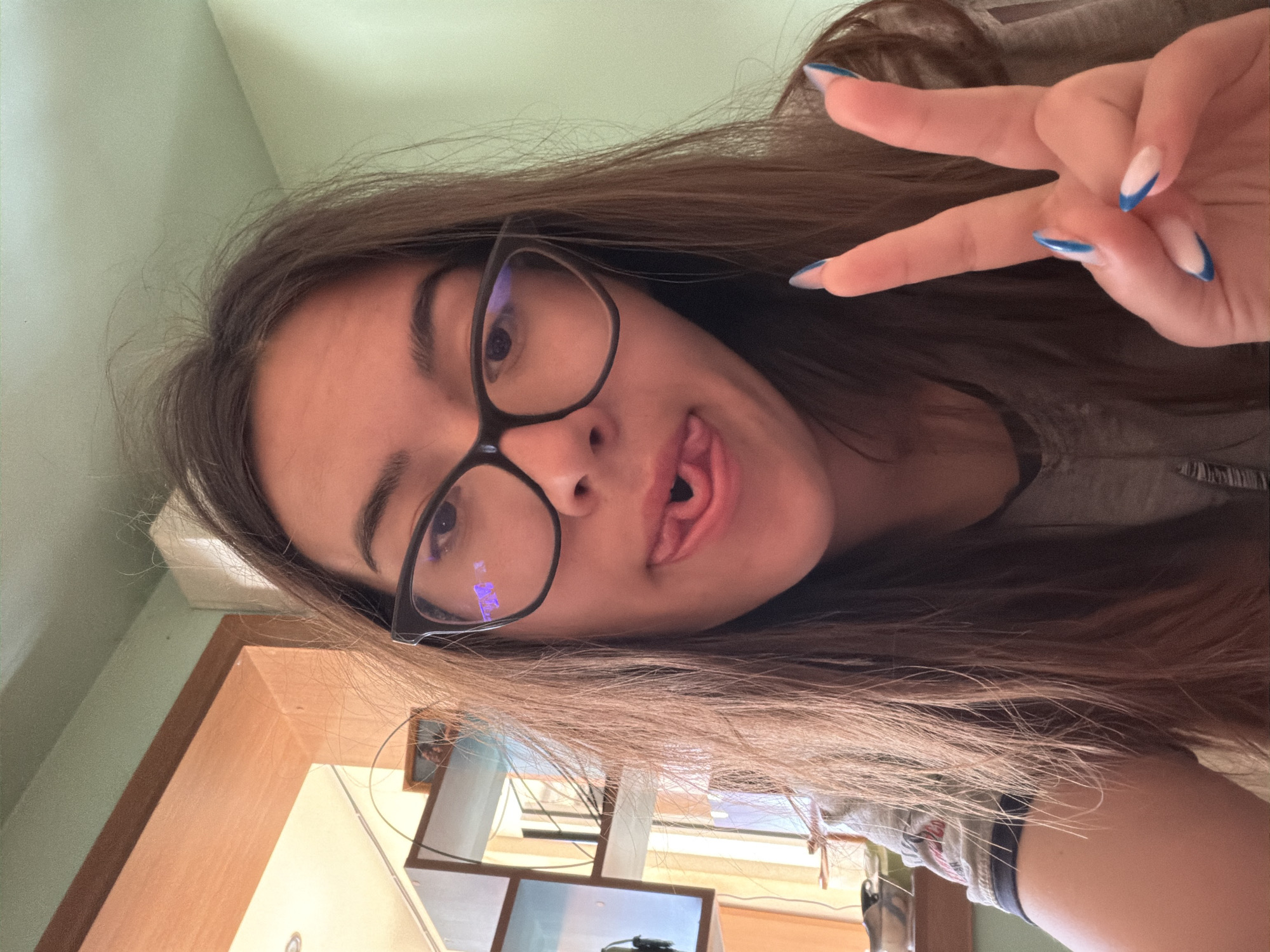 A young woman with light brown hair is taking a selfie indoors. She is wearing black-rimmed glasses and a gray top. She is sticking her tongue out and making a peace sign with her right hand, showing off her manicured nails with a blue design. In the background, there is a wooden frame, a picture on the wall, and a multi-screen setup. The location is suggested to be in Silistra, Bulgaria.